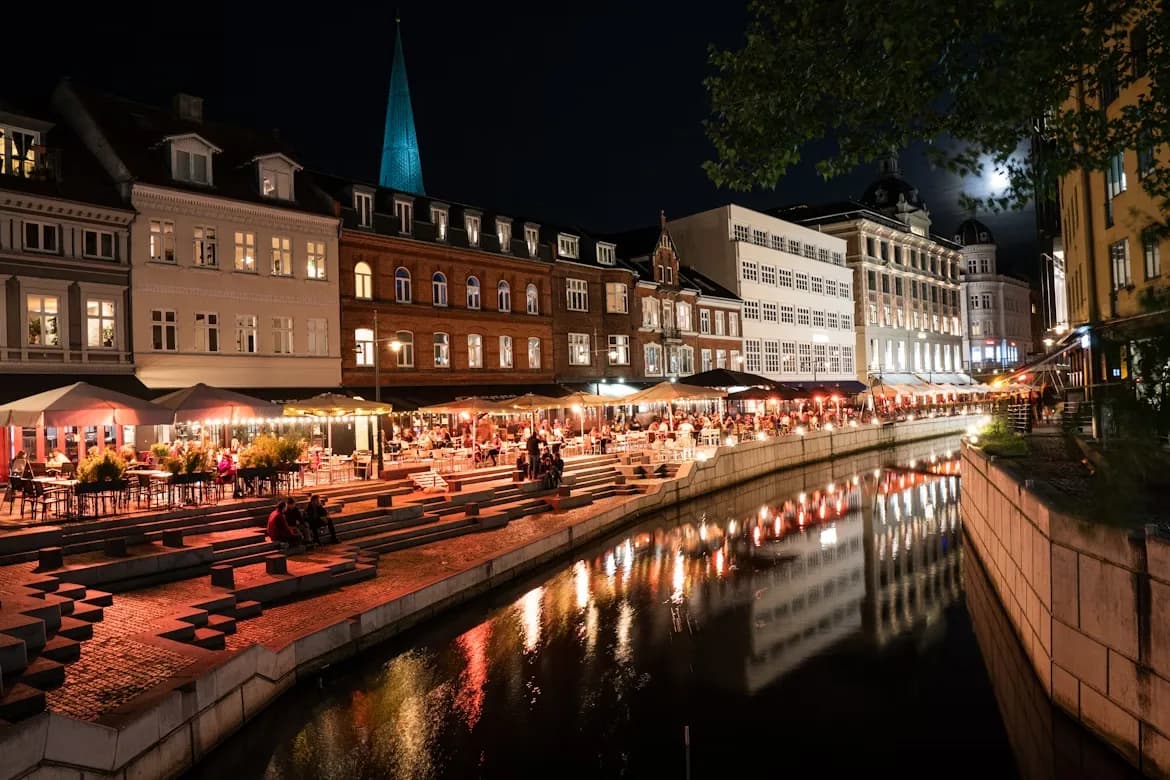 The lively Åboulevarden canal in Aarhus comes alive at night, with people enjoying the vibrant atmosphere from the restaurants and cafes lining the waterfront.