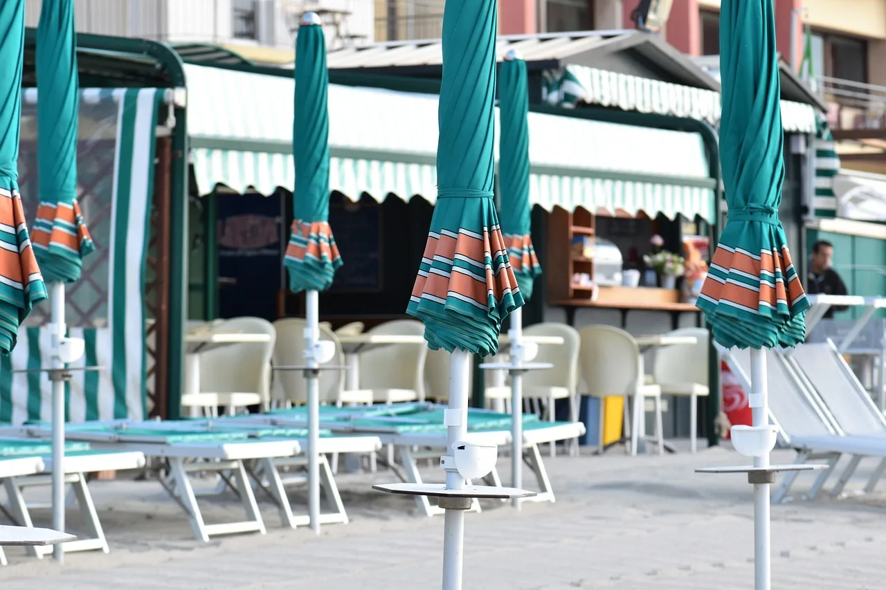 Rows of closed beach umbrellas and empty lounge chairs line a sandy beach, with a cafe and palm trees in the background.