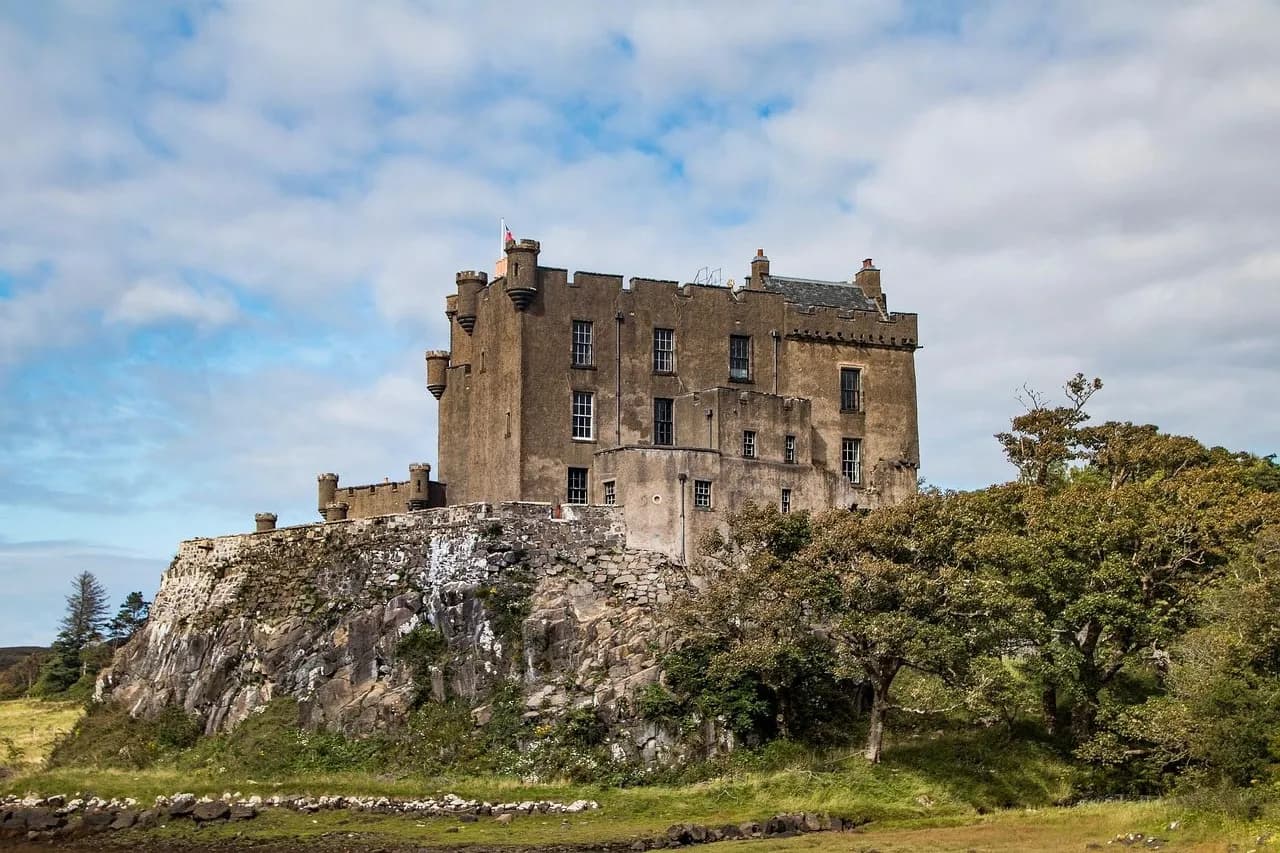 The majestic Dunvegan Castle, a historic fortress with a square tower, stands on a rocky outcrop surrounded by green trees and fields.