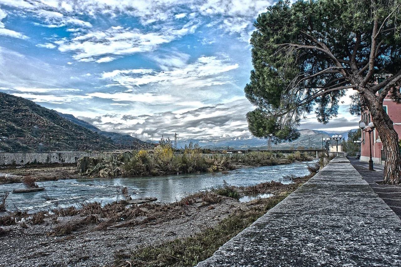 A wide, shallow river flows through a valley, with mountains and trees in the background under a cloudy sky.