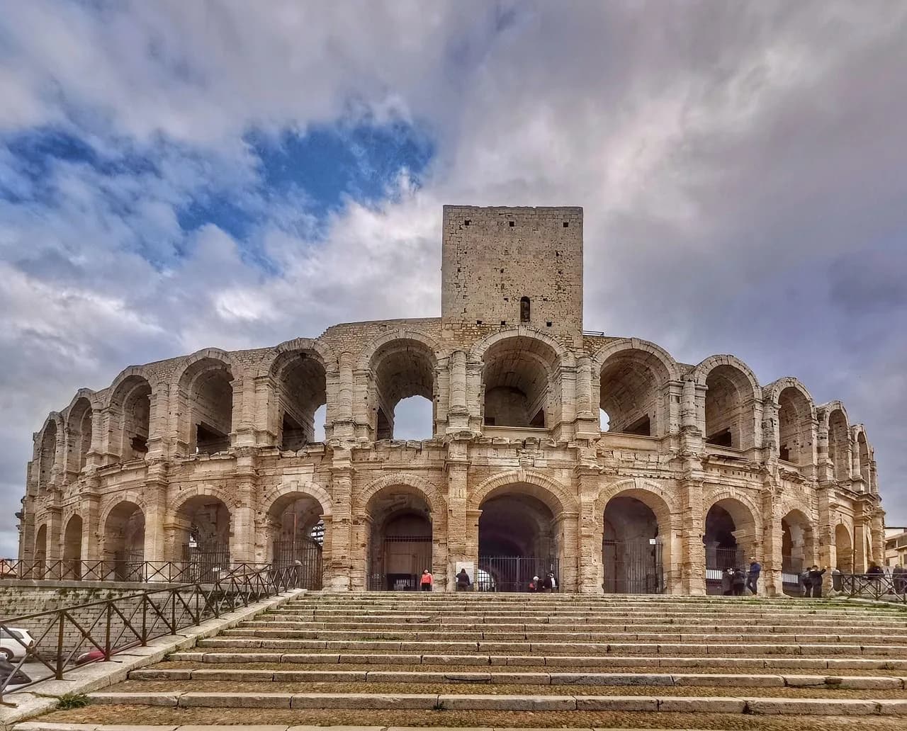 The Roman Amphitheatre of Arles, a well-preserved ancient arena, stands under a cloudy sky.