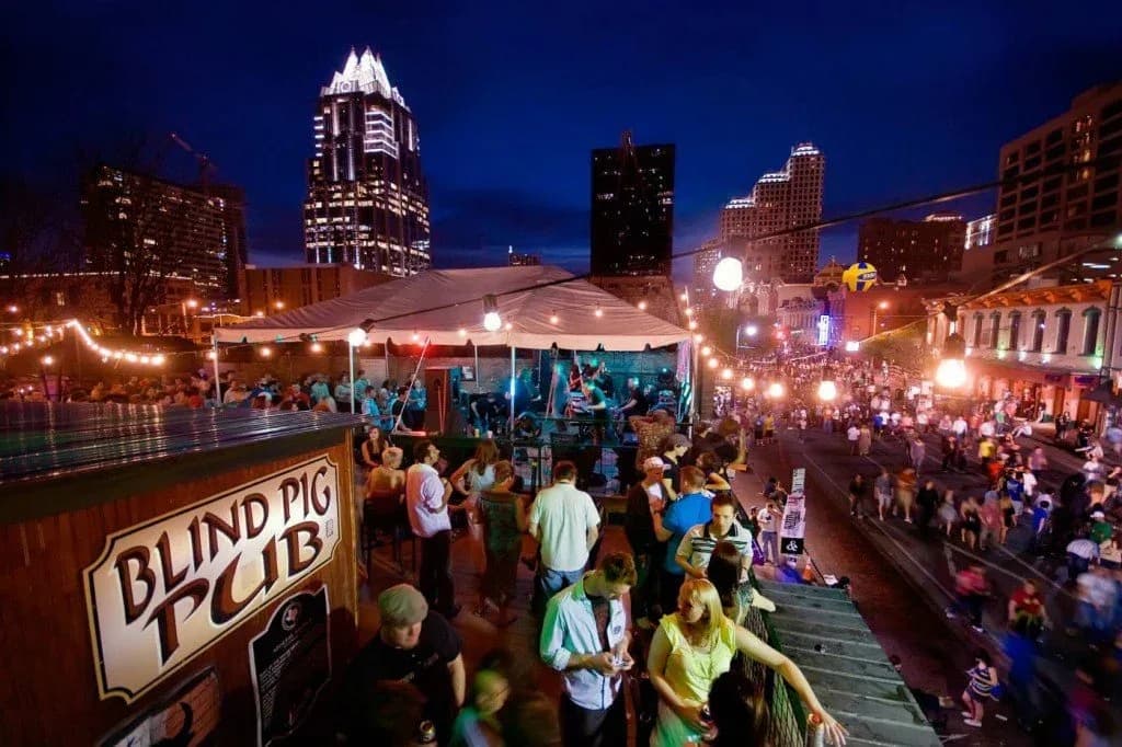 A lively outdoor terrace of a pub is bustling with people at night, with the illuminated Austin skyline in the background.