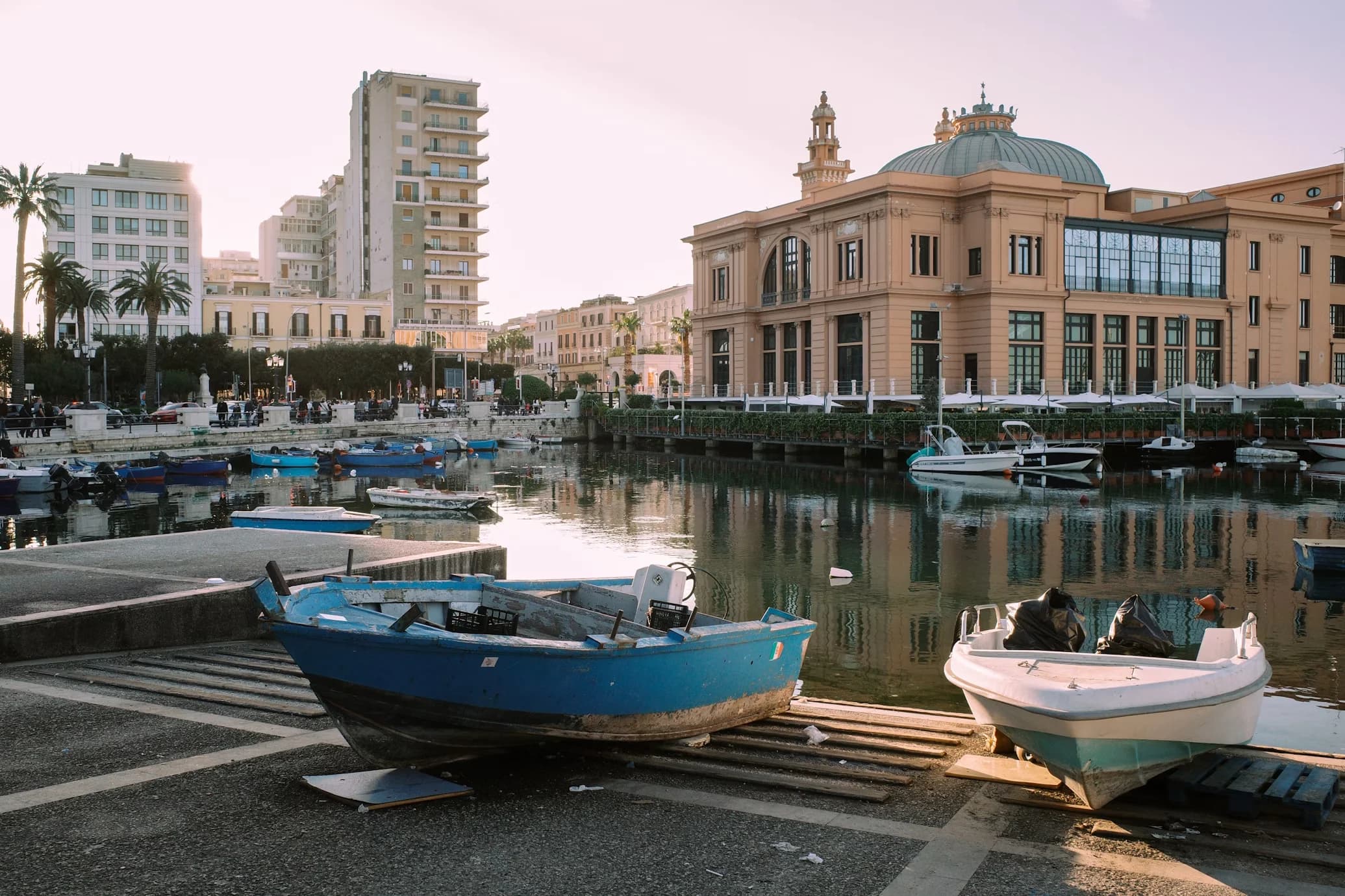 Small fishing boats are pulled up on a wooden ramp, with a grand, historic palace and palm trees lining the harbor in Bari.