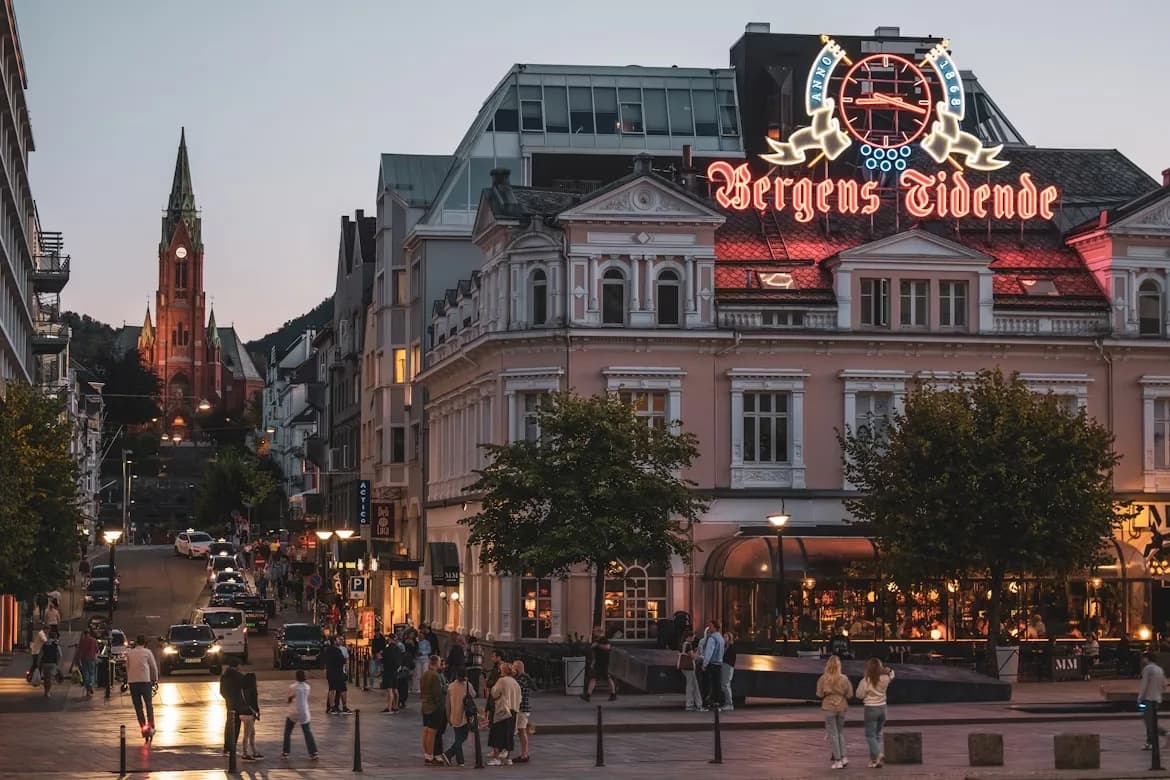 A busy street in Bergen is illuminated by streetlights and neon signs at dusk, with a church steeple and mountains in the background.