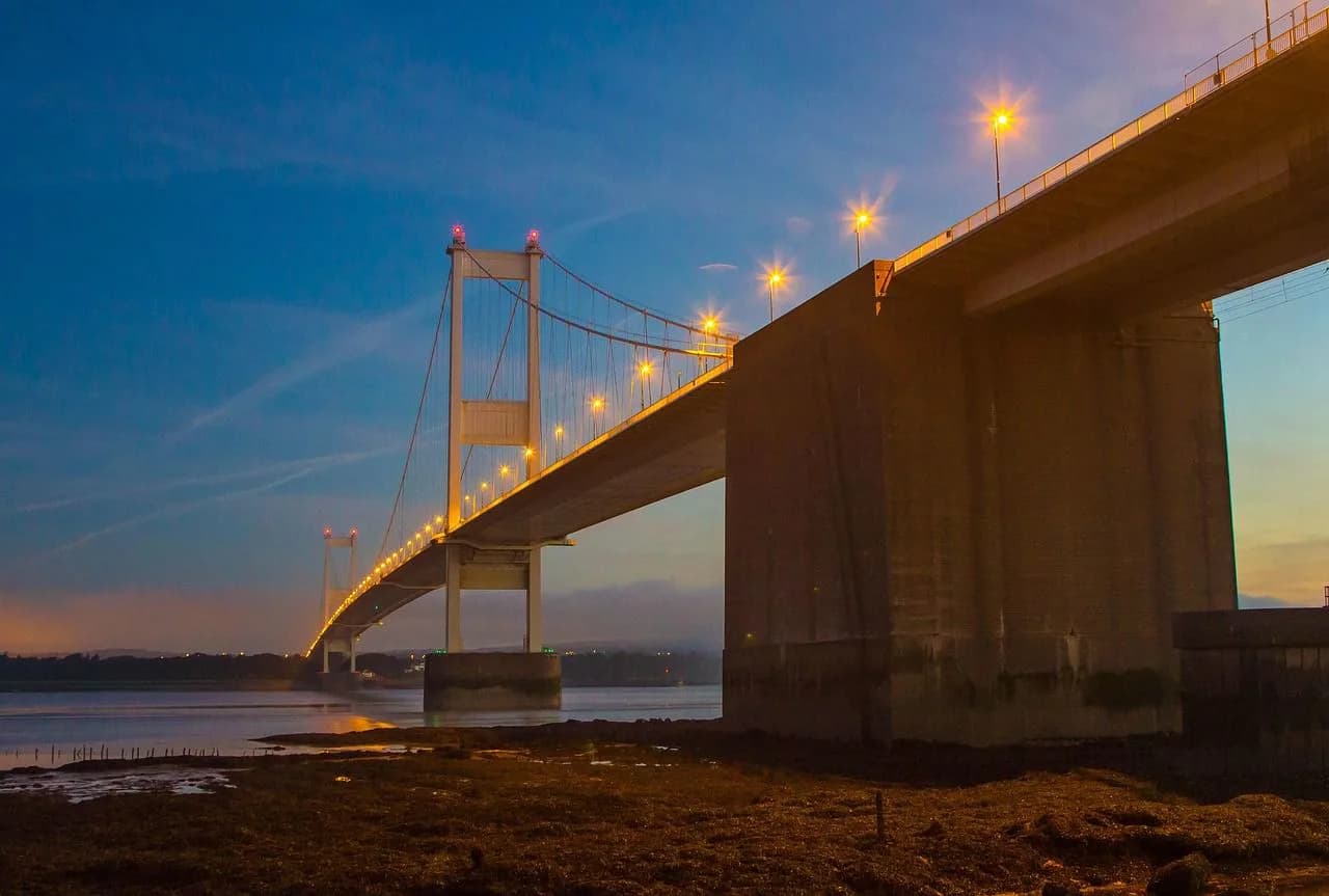 The Severn Bridge, a massive suspension bridge with illuminated supports, connects two shorelines at night.