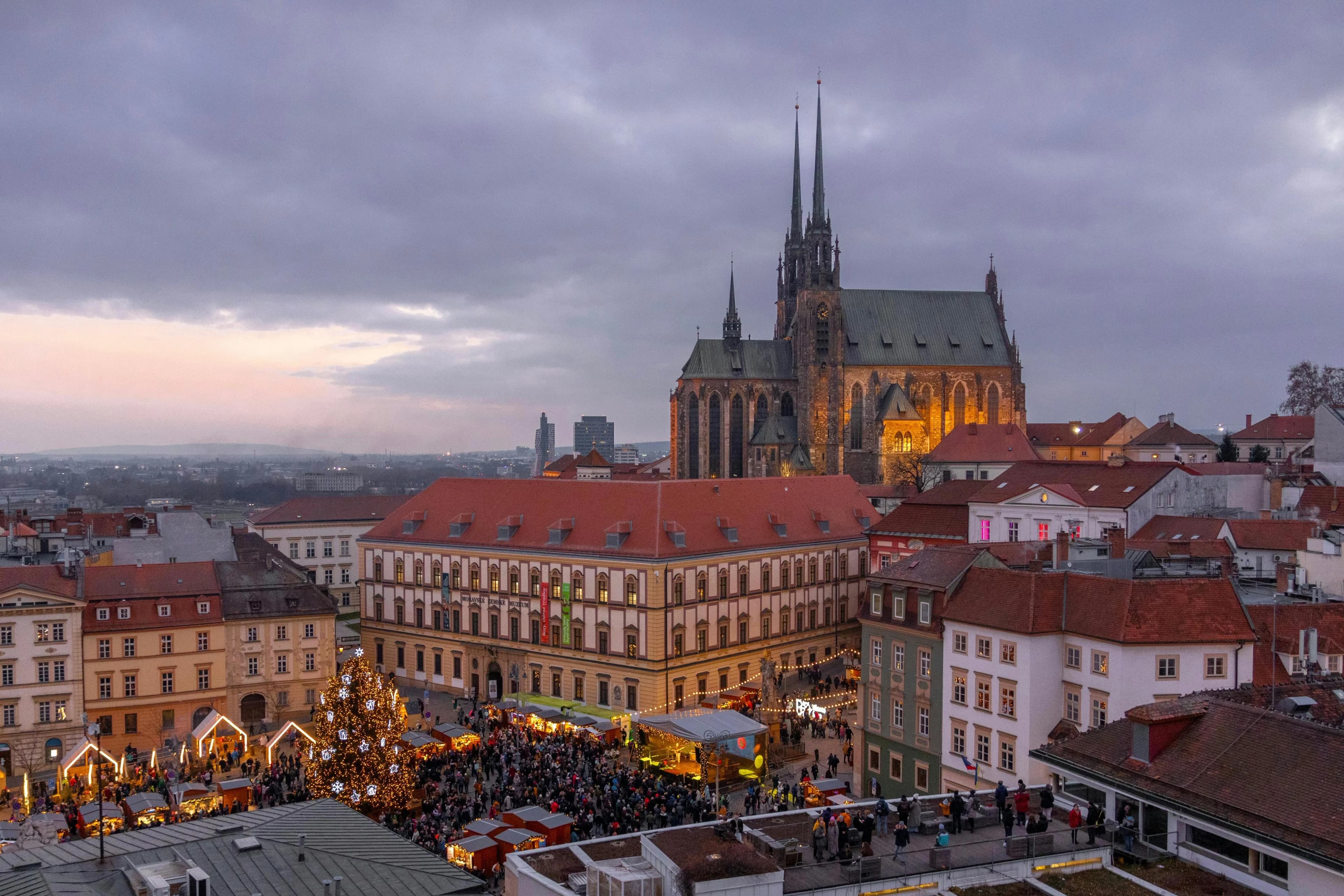 The historic city of Brno is beautifully illuminated at dusk, with a festive Christmas tree and a bustling market in the square below.