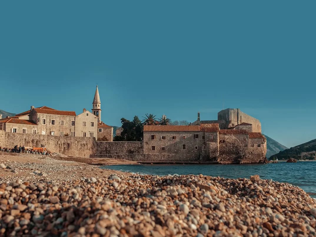 The fortified walls and buildings of Budva's Old Town are nestled beside a tranquil beach with small pebbles.
