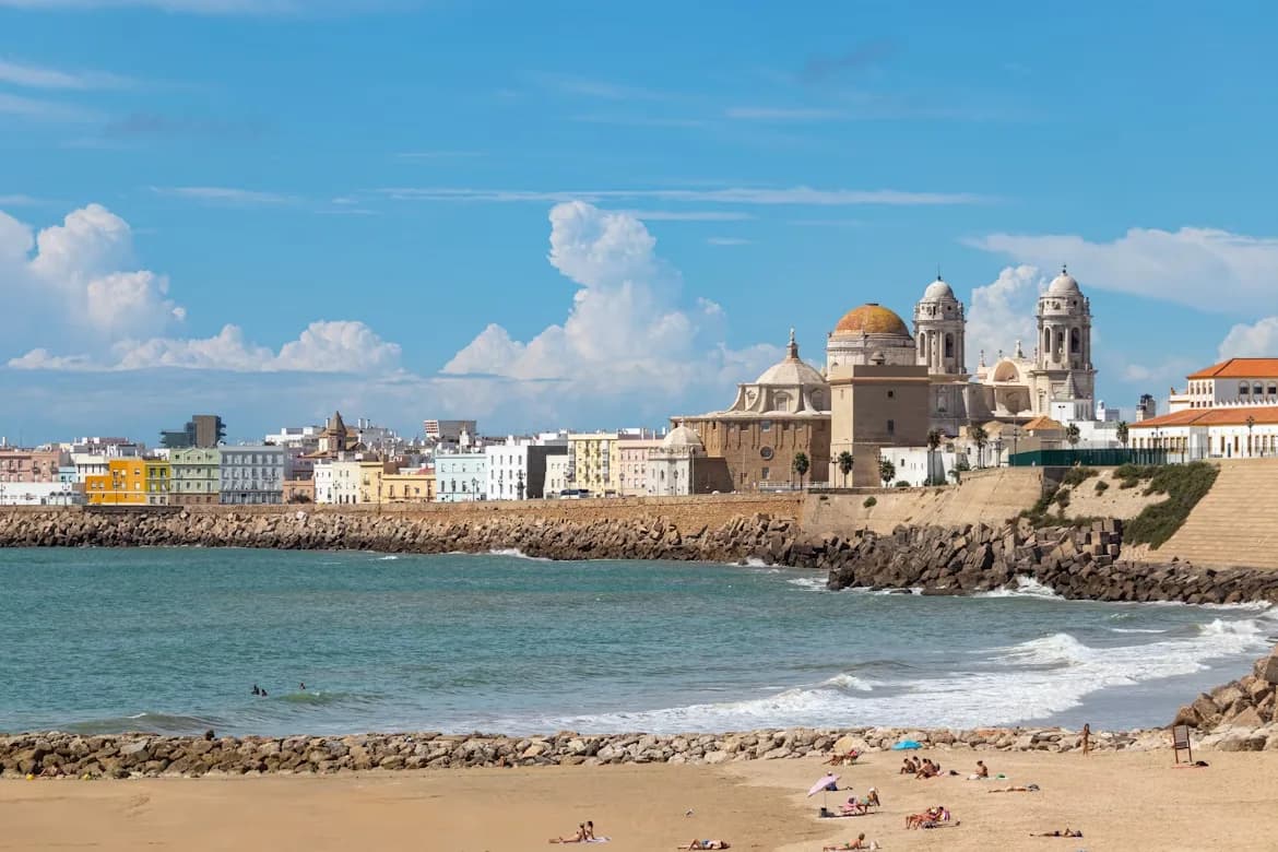 People relax on a sandy beach, with waves crashing on the shore and the historic Cádiz Cathedral in the background.