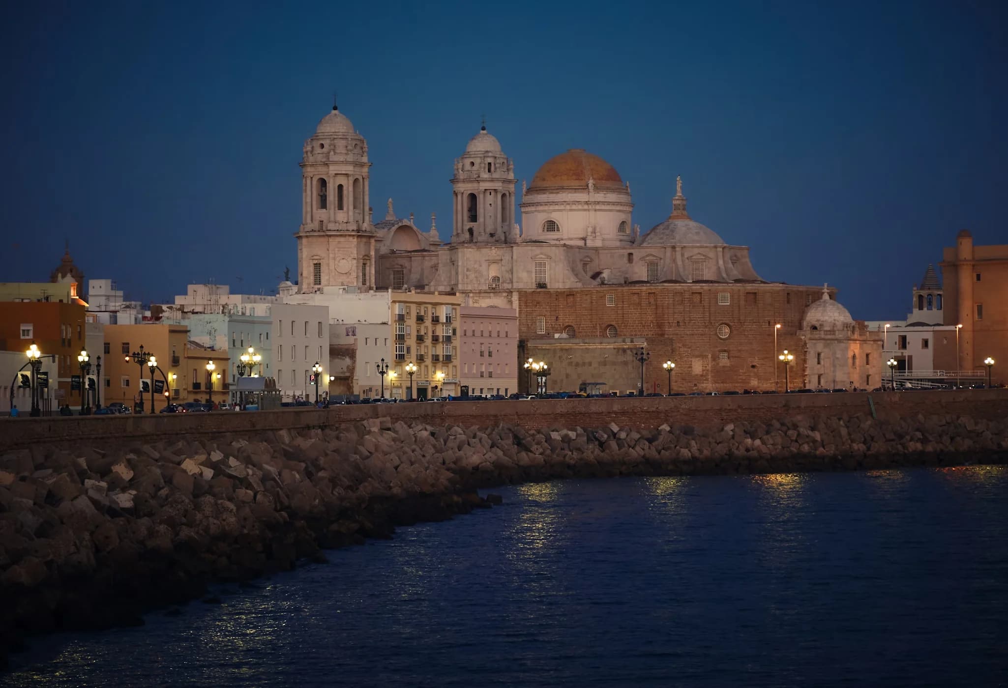 The illuminated Cádiz Cathedral is viewed from across the water at dusk, its lights reflecting on the tranquil sea.