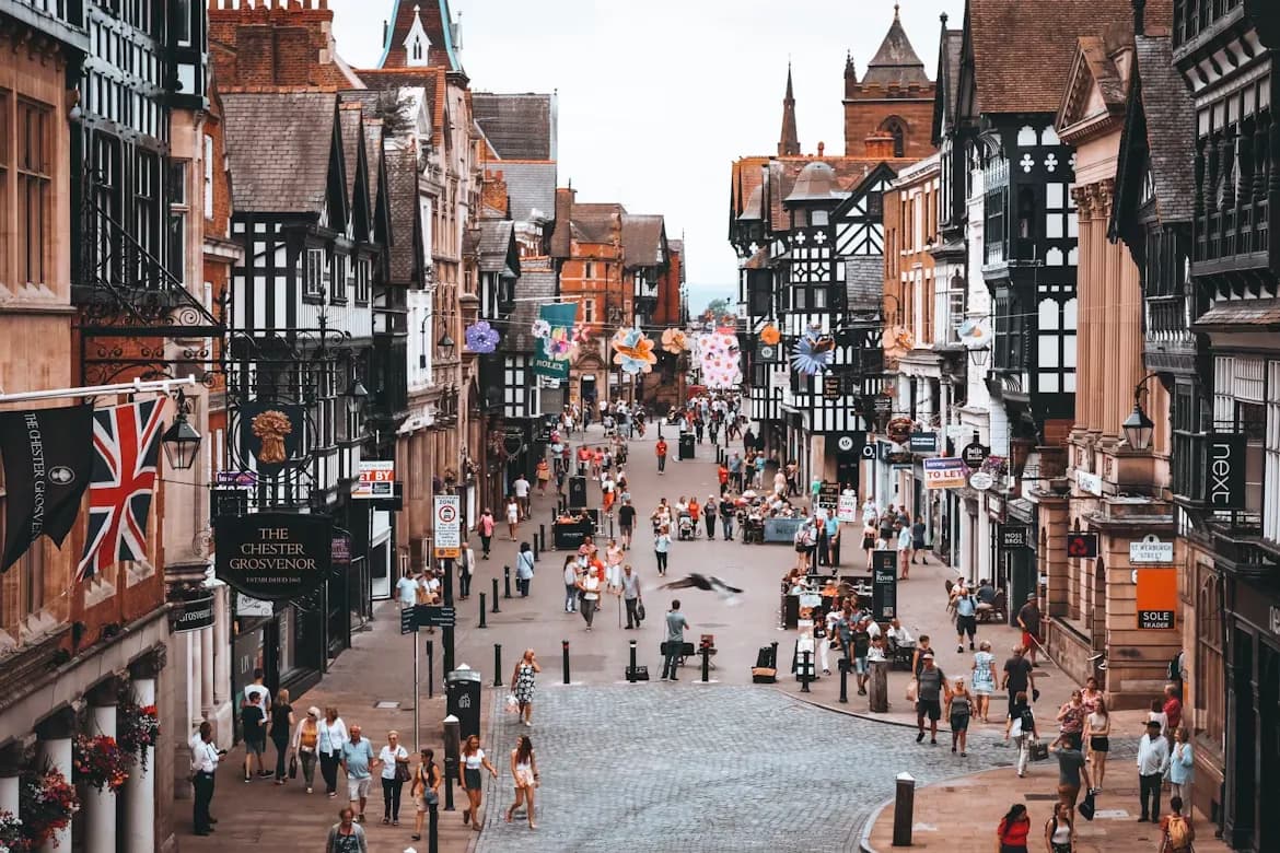 People walk along a historic shopping street in Chester, with the unique black and white timber-framed buildings and a clock tower in the background.