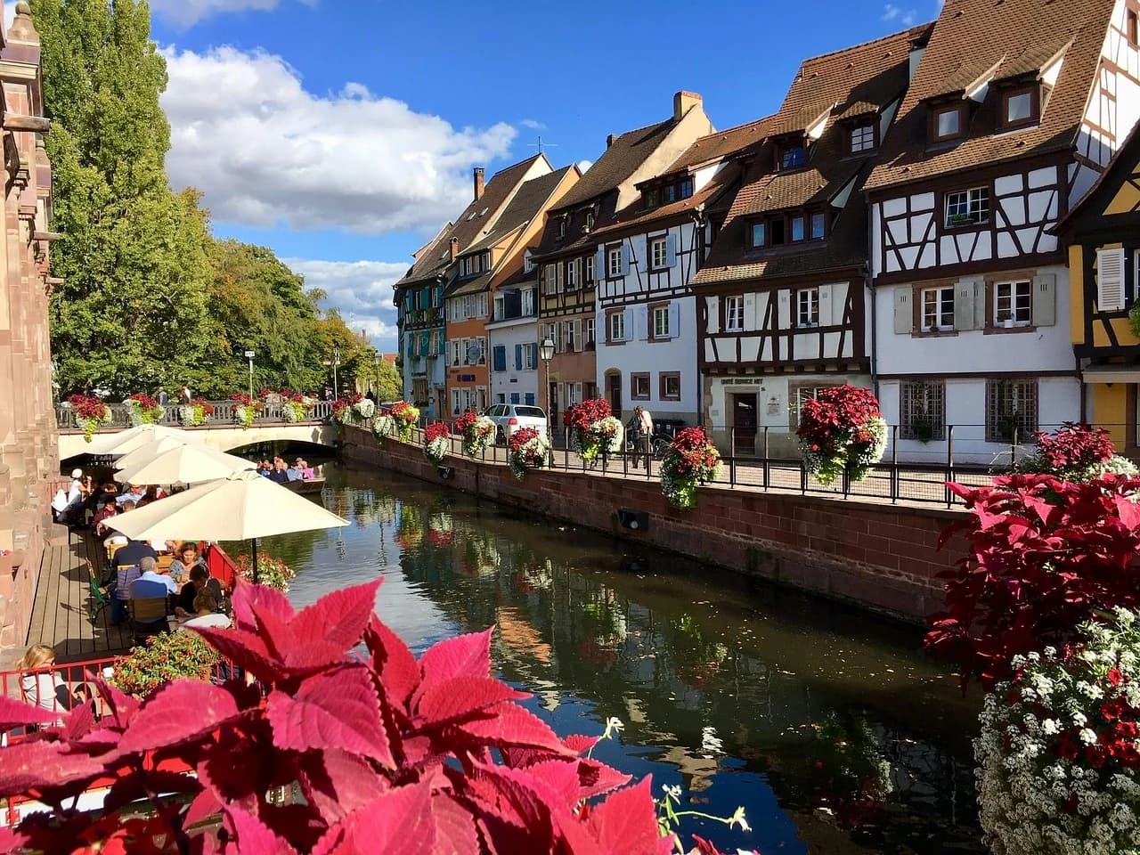 Outdoor cafes and beautiful red flowers line the canal in the "Little Venice" district of Colmar, with historic buildings in the background.