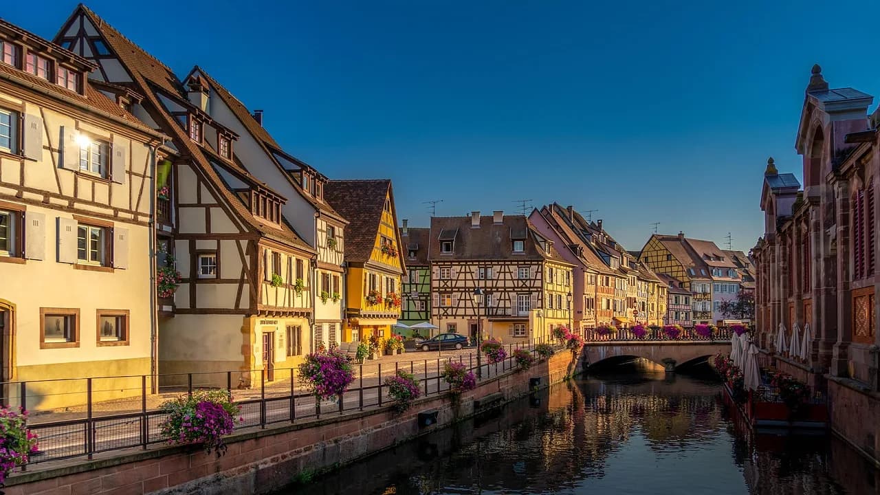 The beautiful half-timbered houses of Colmar are reflected in a tranquil canal under a clear blue sky.