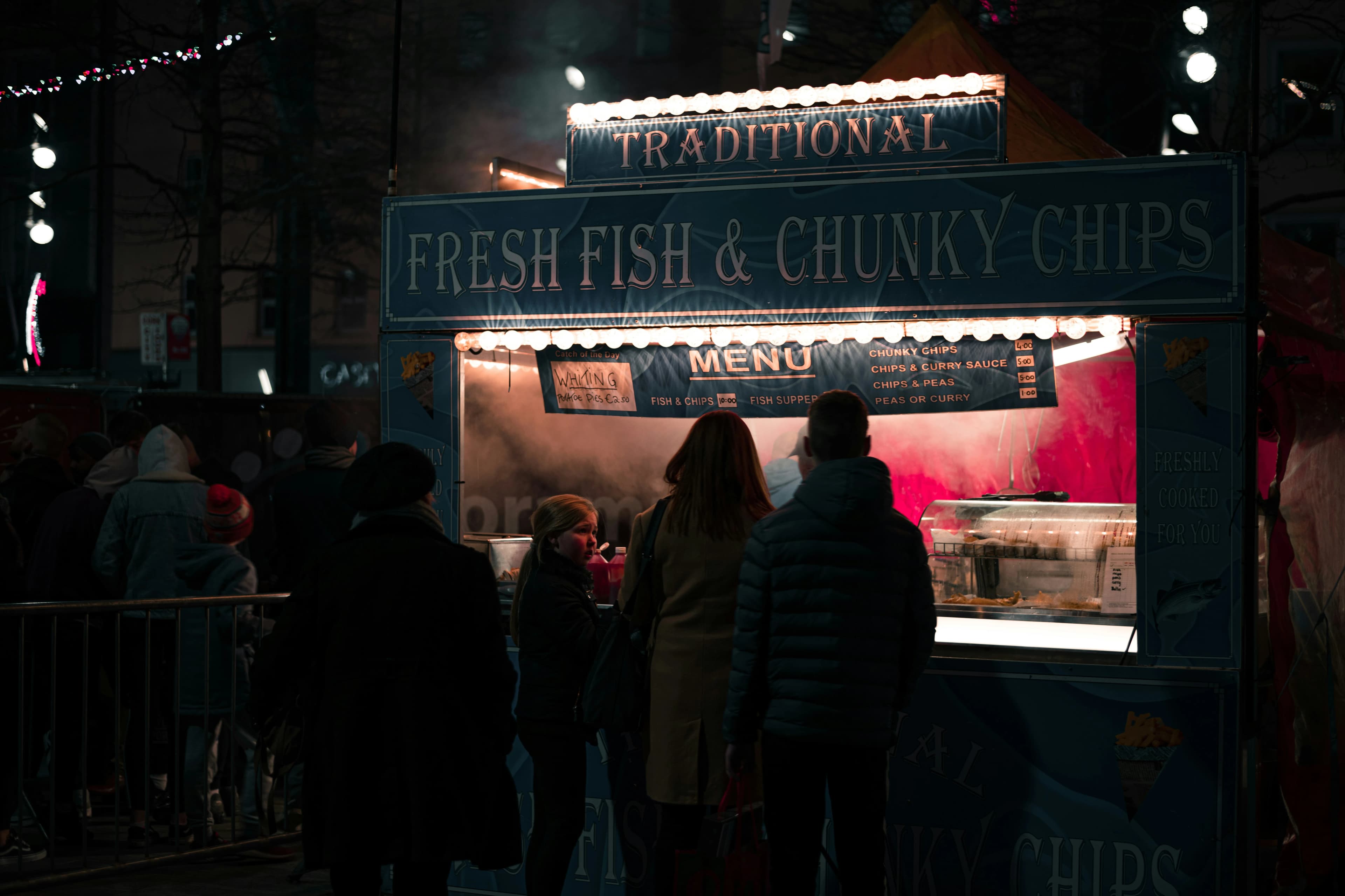 People line up at a traditional fish and chips food stall, with glowing lights and a menu advertising the local specialty.