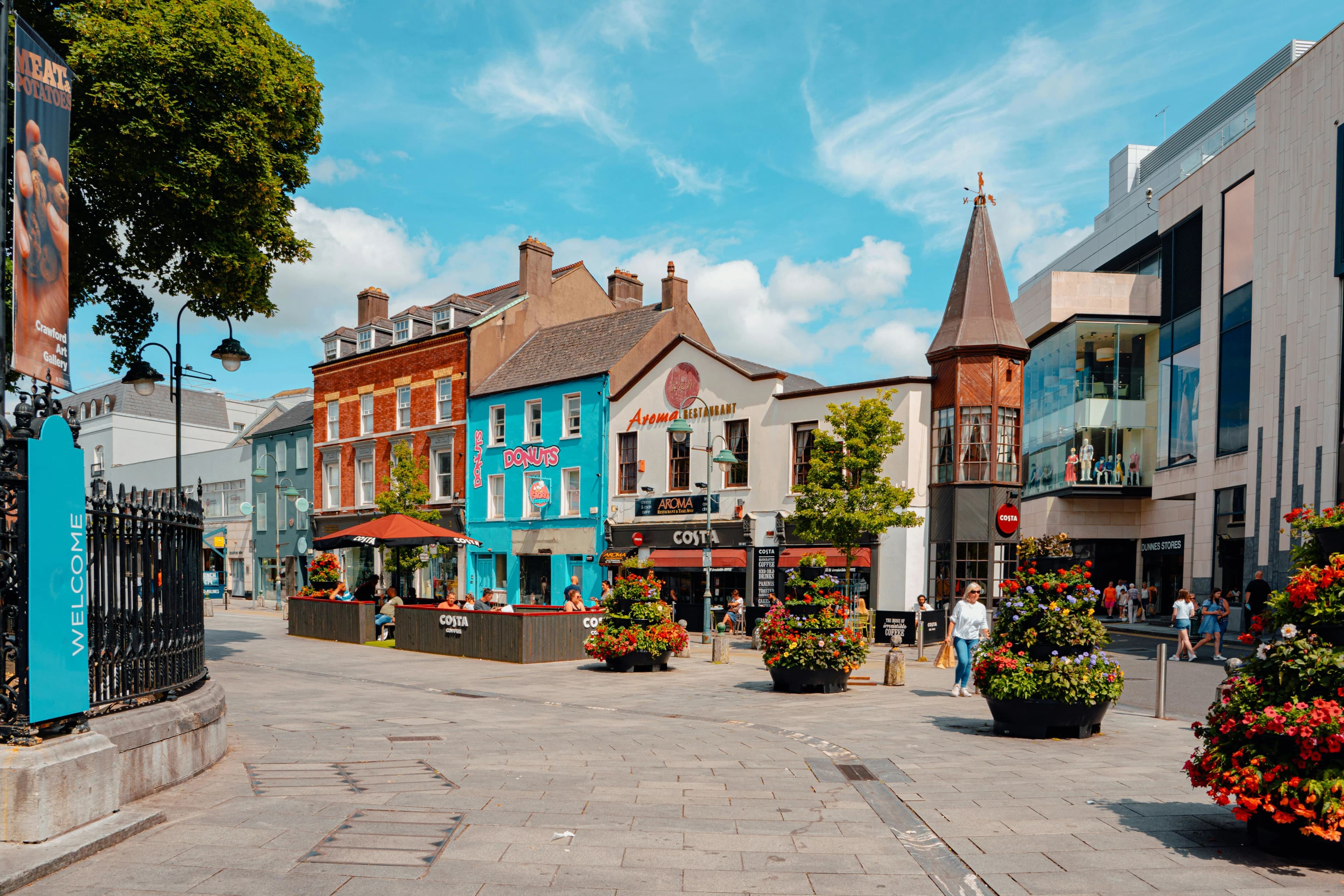 The vibrant streets of Cork are bustling with people, with colorful storefronts and overflowing flower baskets adding to the lively atmosphere.