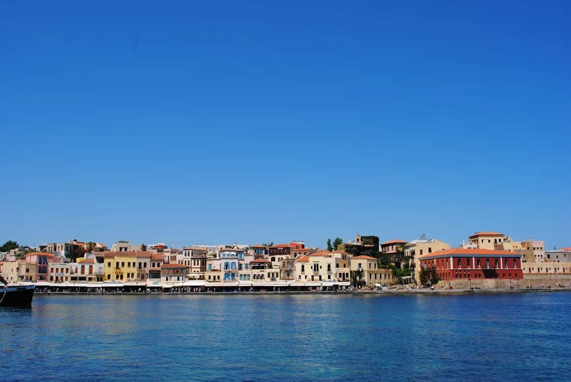 The colorful, historic buildings of the Venetian harbor of Chania line the waterfront under a clear blue sky.