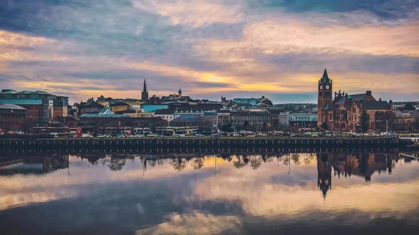 The city of Derry is beautifully reflected in the calm waters of the River Foyle, with the historic Guildhall and its clock tower in the foreground.