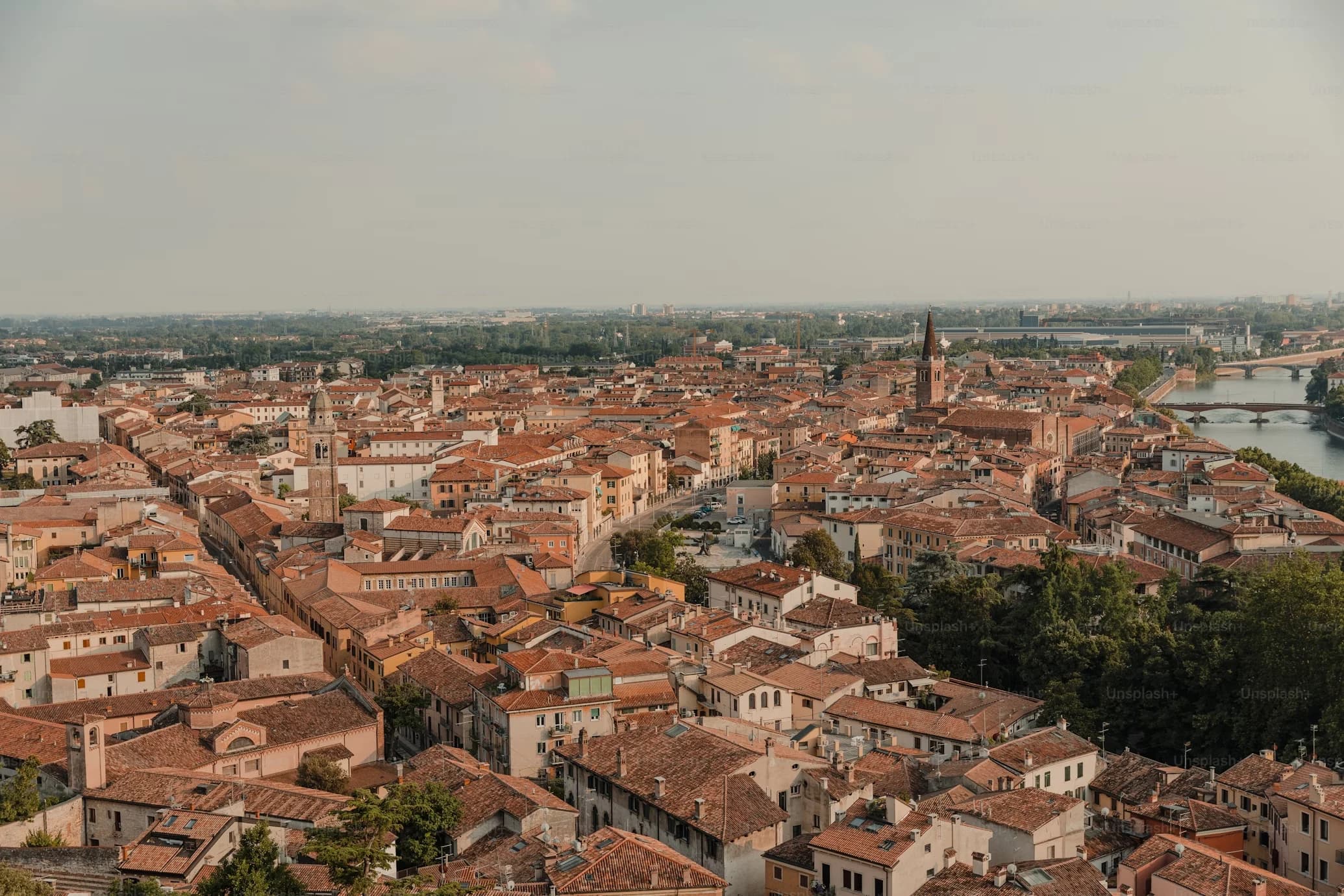 A wide view of a European city shows a sea of red-tiled roofs, with a river and a bridge in the distance.