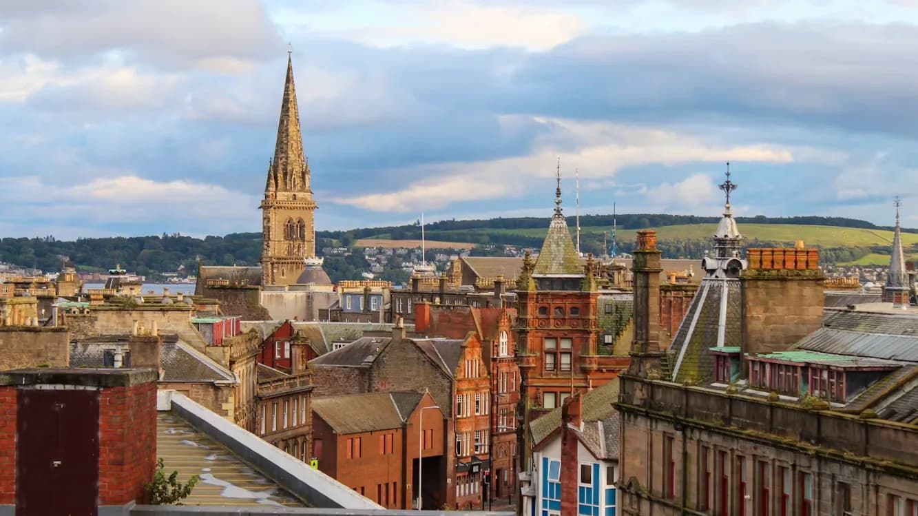 The skyline of Dundee features a striking gothic spire rising above the rooftops, with rolling green hills in the distance.