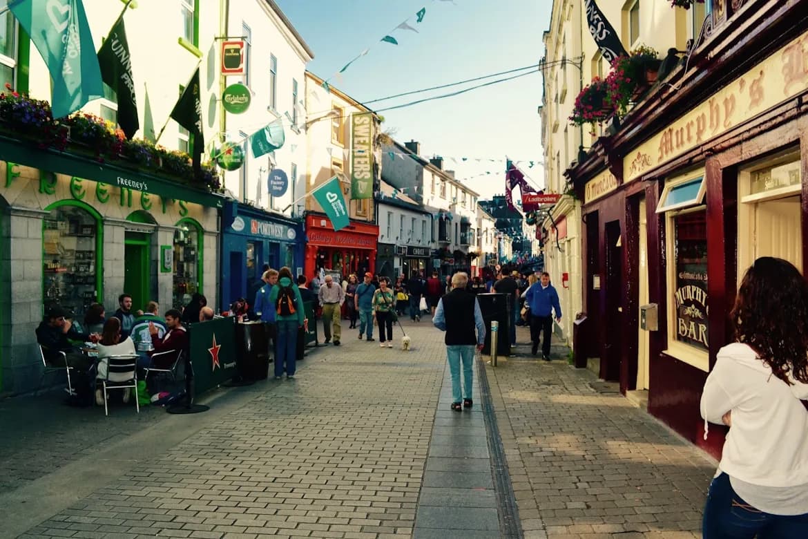 A bustling street in Galway is filled with people and colorful pub facades, showcasing the city's lively atmosphere.