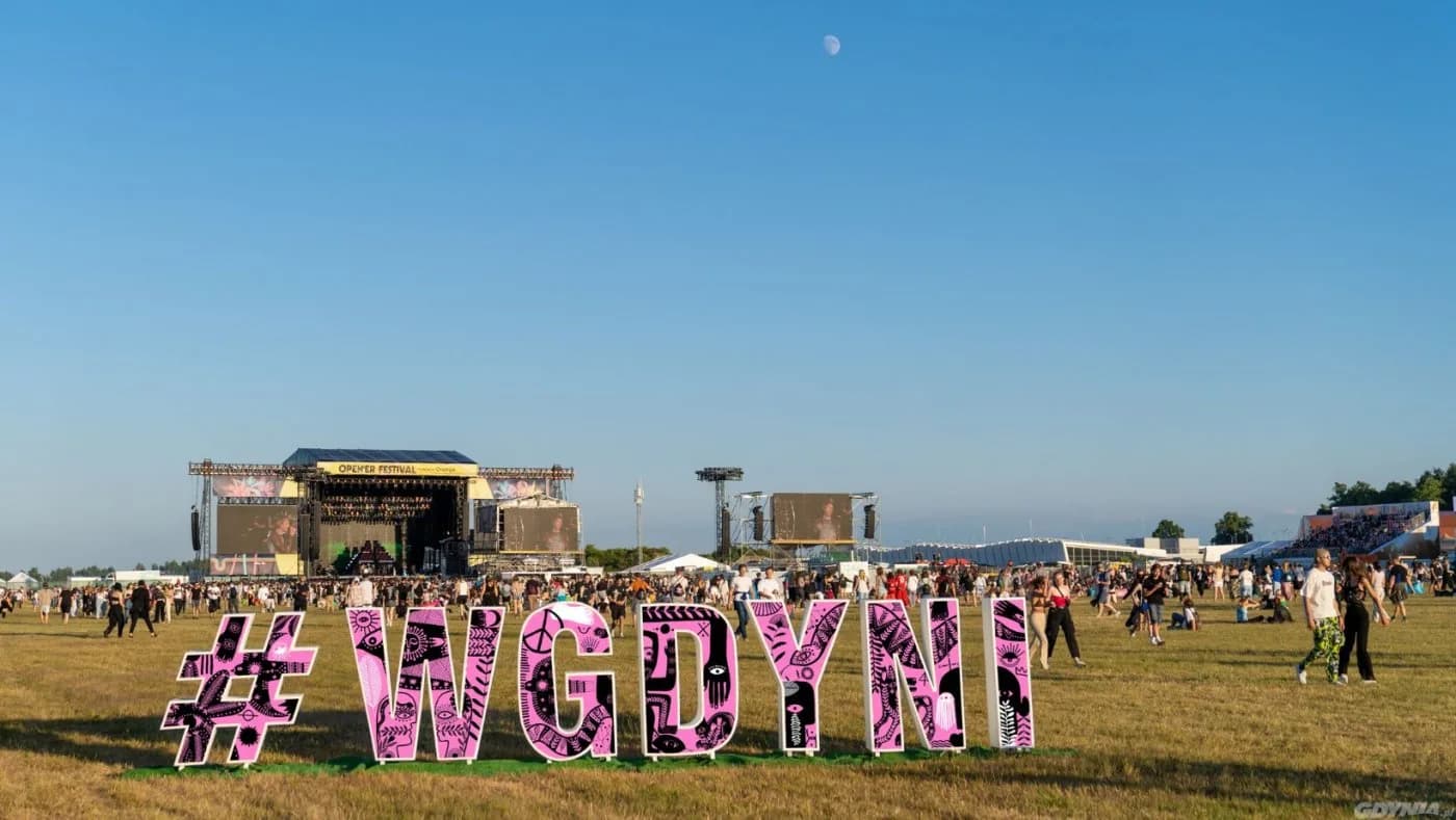A large, decorated sign for the Open'er Festival stands on a grassy field, with a stage and a crowd of people in the background.