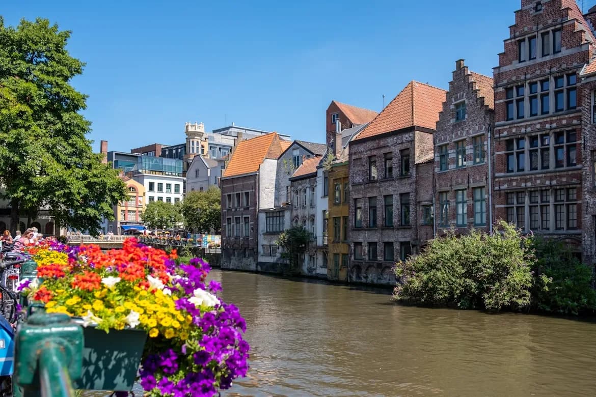 A tranquil canal runs through Ghent, with colorful flowers and historic, gabled buildings lining the banks.
