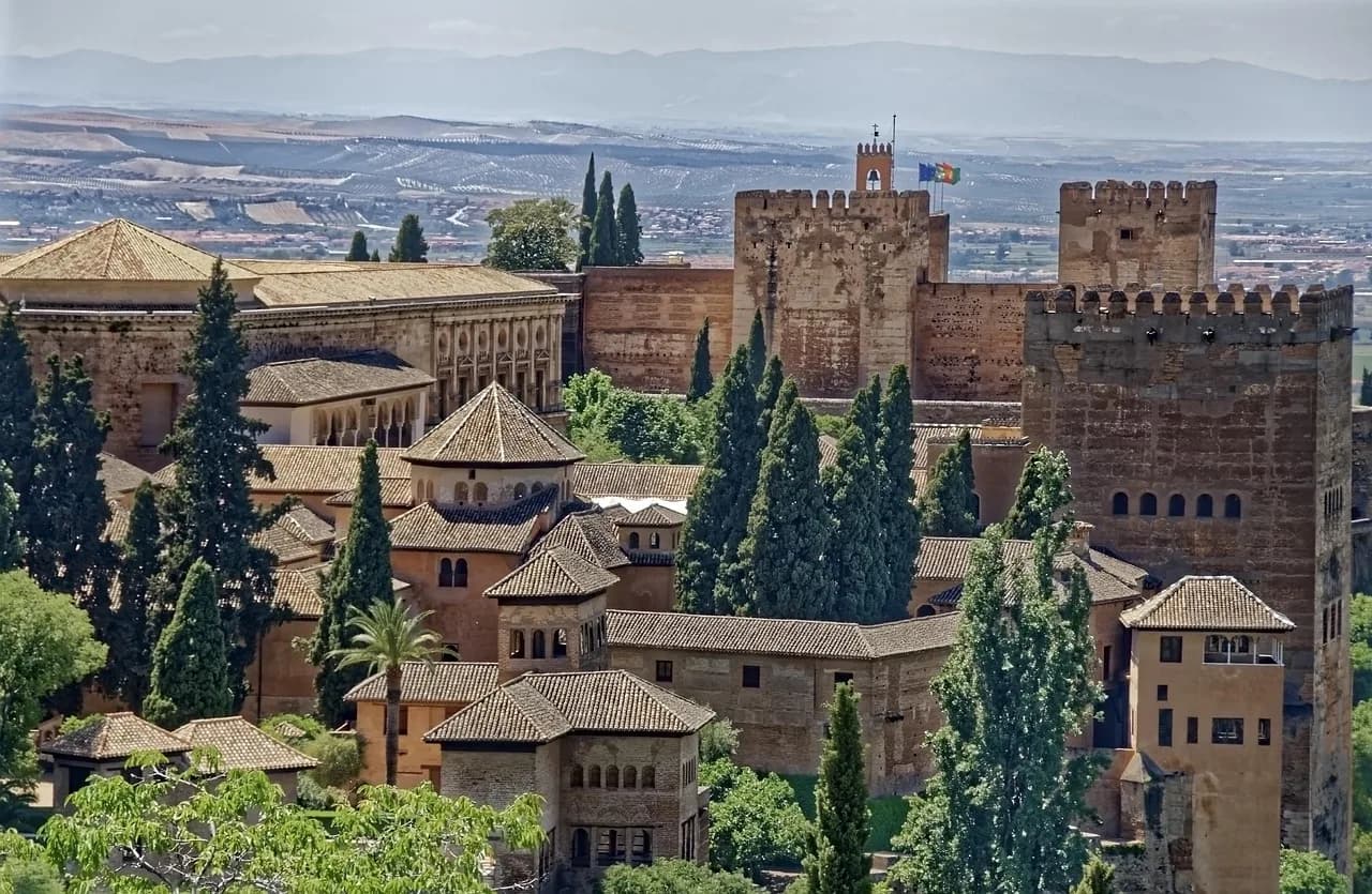The fortified walls and towers of the Alcazaba, part of the Alhambra, are framed by lush greenery, with the city of Granada visible in the distance.