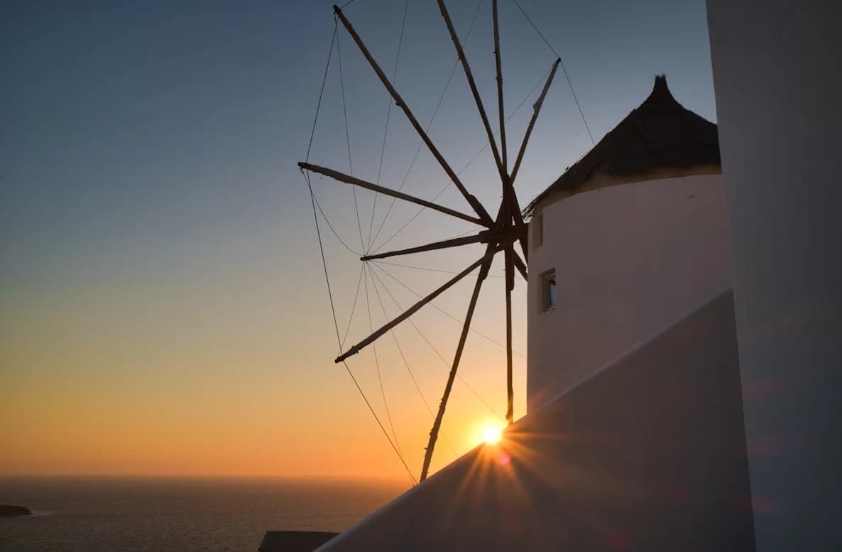 A traditional Greek windmill with large sails is silhouetted against a vibrant sunset over the sea.