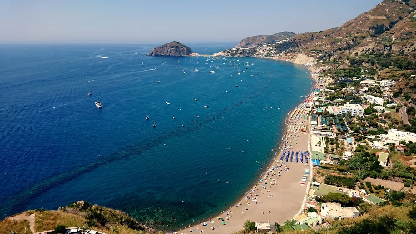 A stunning aerial view shows the coastline of Ischia, with its sandy beaches, sparkling blue waters, and a scattering of boats.