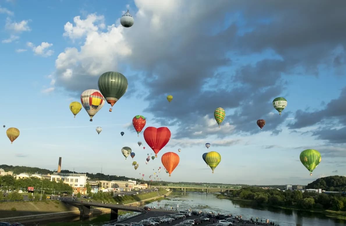A multitude of colorful hot air balloons float over the city of Kaunas, with the river and bridges below.