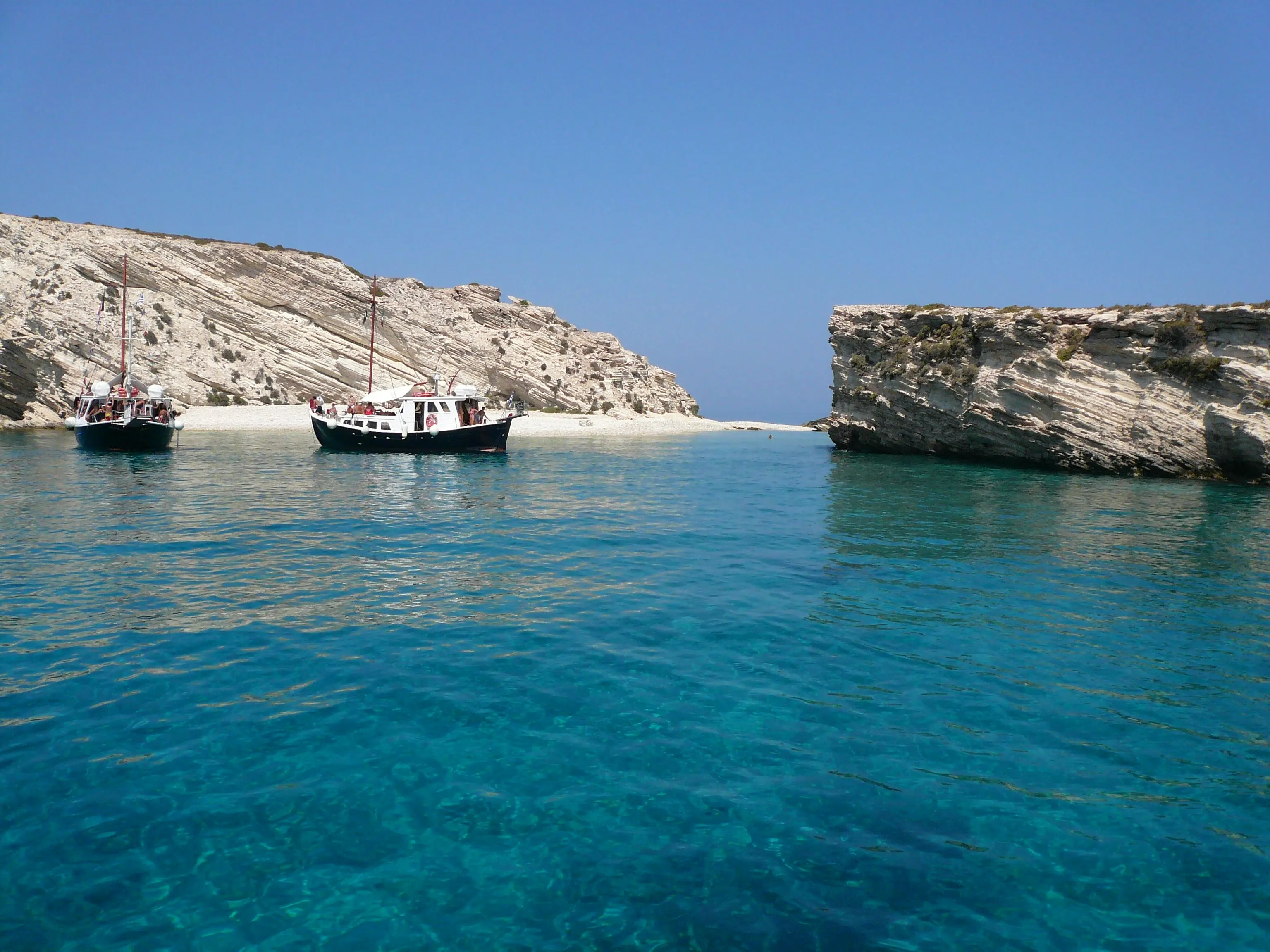 Two boats are anchored in a secluded cove with stunning turquoise water, framed by impressive rocky cliffs.