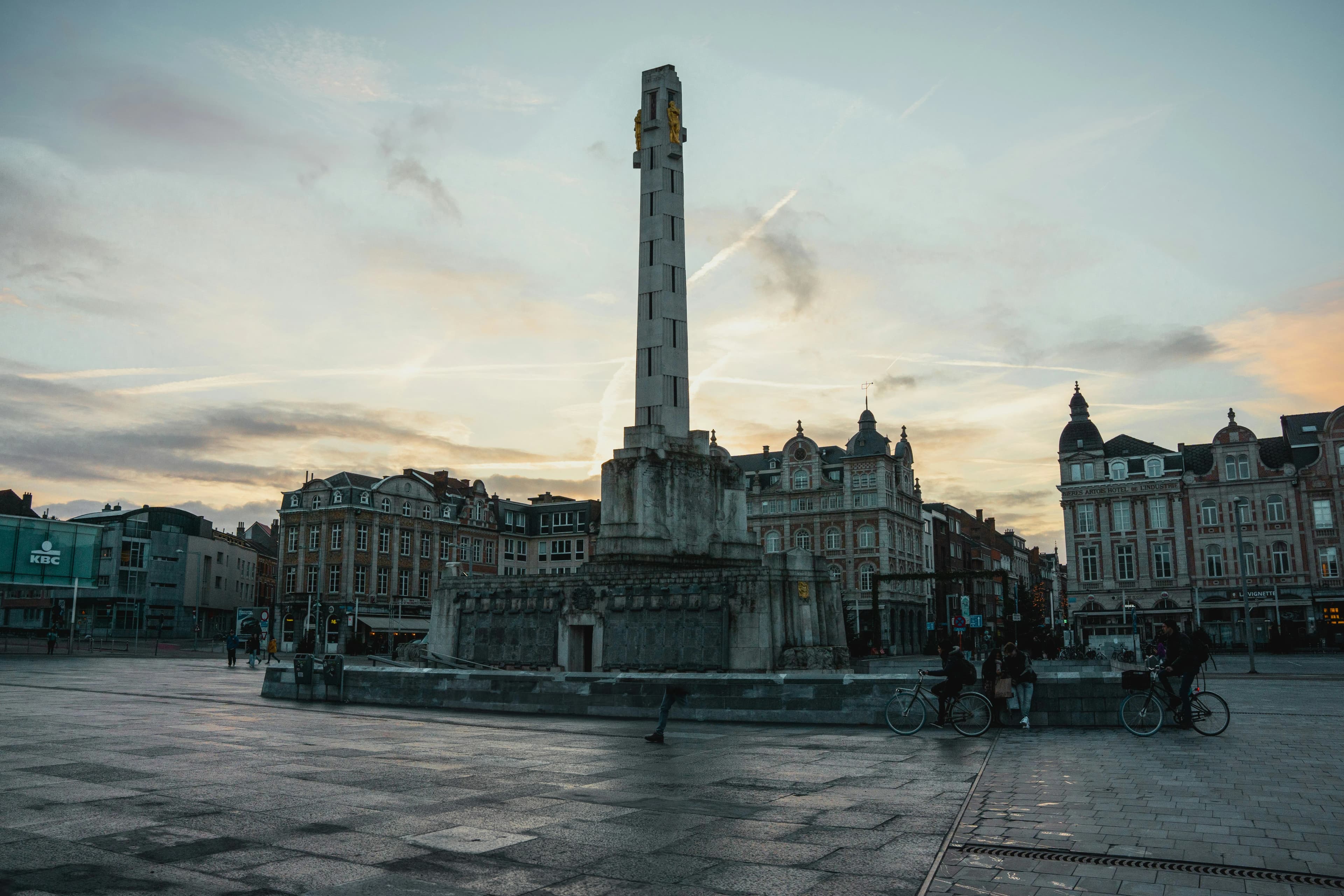 A monument stands in the center of a wide, empty square in Leuven, surrounded by historic buildings and an evening sky.