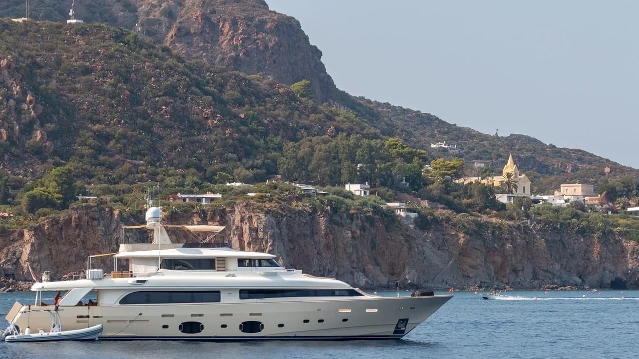 A large, luxurious yacht is anchored in a bay, with a dramatic, rocky coastline and a church steeple on a hill.