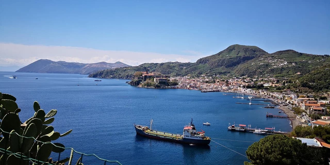 A small cargo ship is moored in a beautiful bay, with the historic town of Lipari and a green hillside in the background.