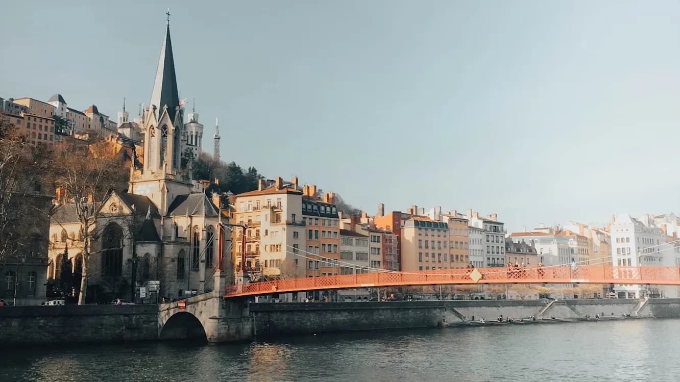 A historic church with a soaring steeple is perched on a hillside, overlooking a red bridge that spans the Saône River.