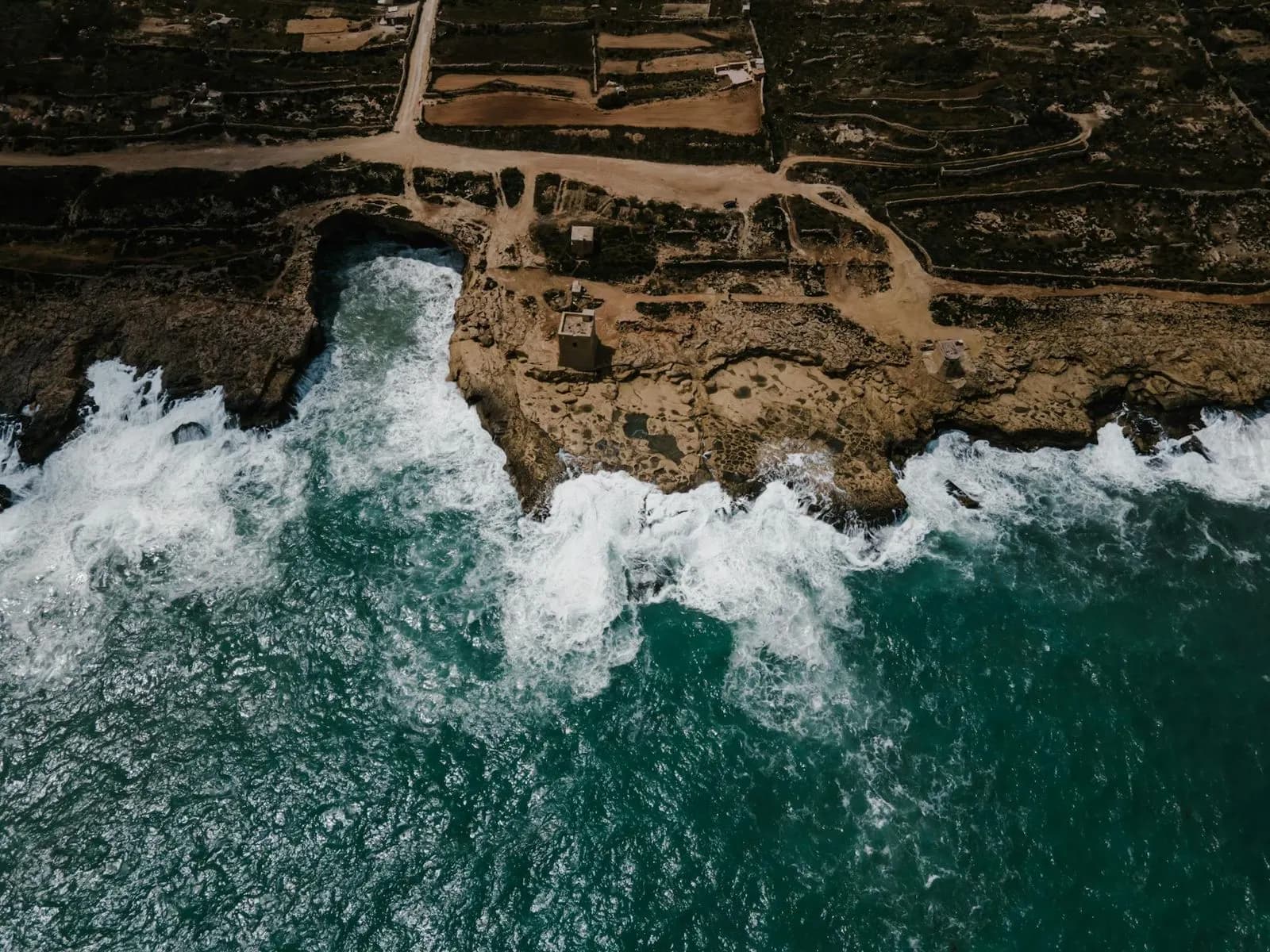 An aerial view captures the dramatic, rocky coastline of Malta, with waves crashing against the shore and a historic watchtower.