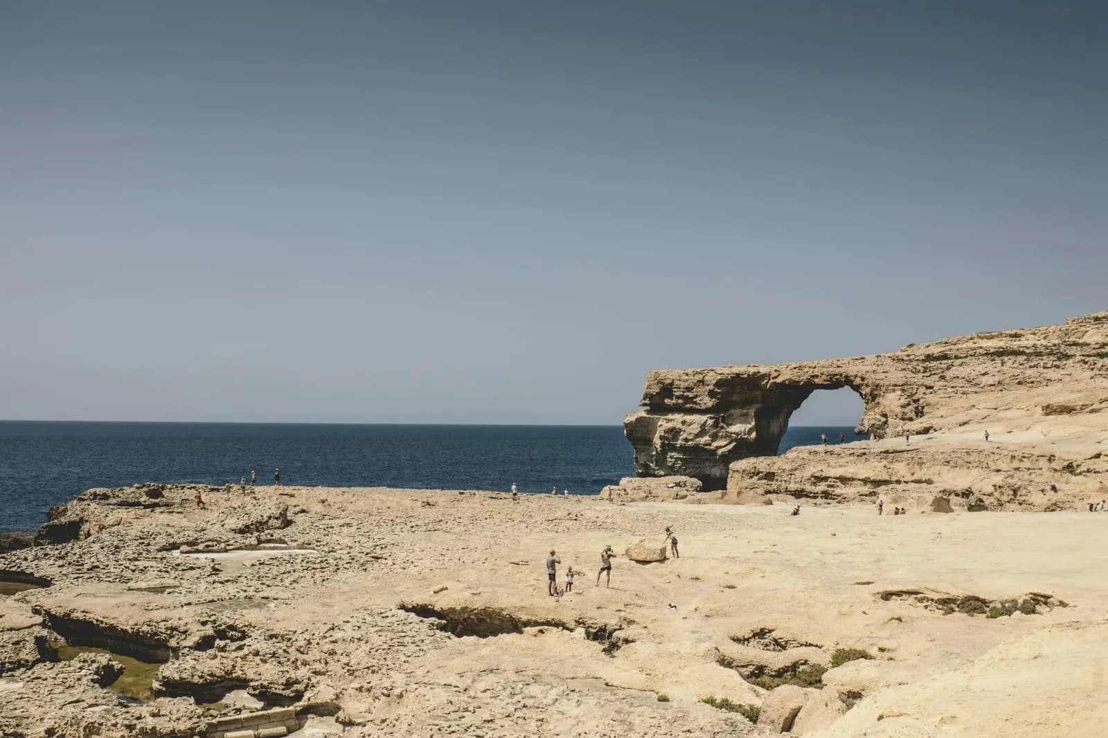 A large natural archway in a rock formation, a former landmark of Malta, is viewed from a dry, rocky outcrop.