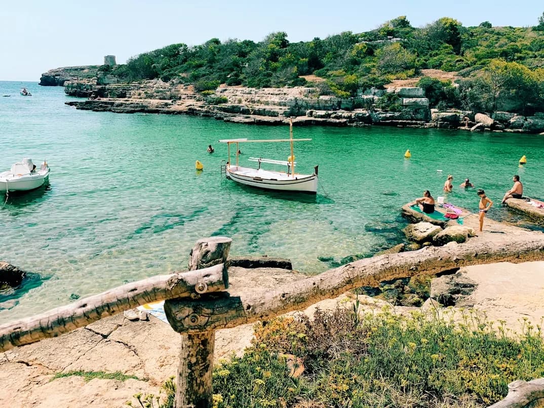 A secluded bay with clear, green water is filled with boats and people swimming, surrounded by a rocky coastline and green trees.