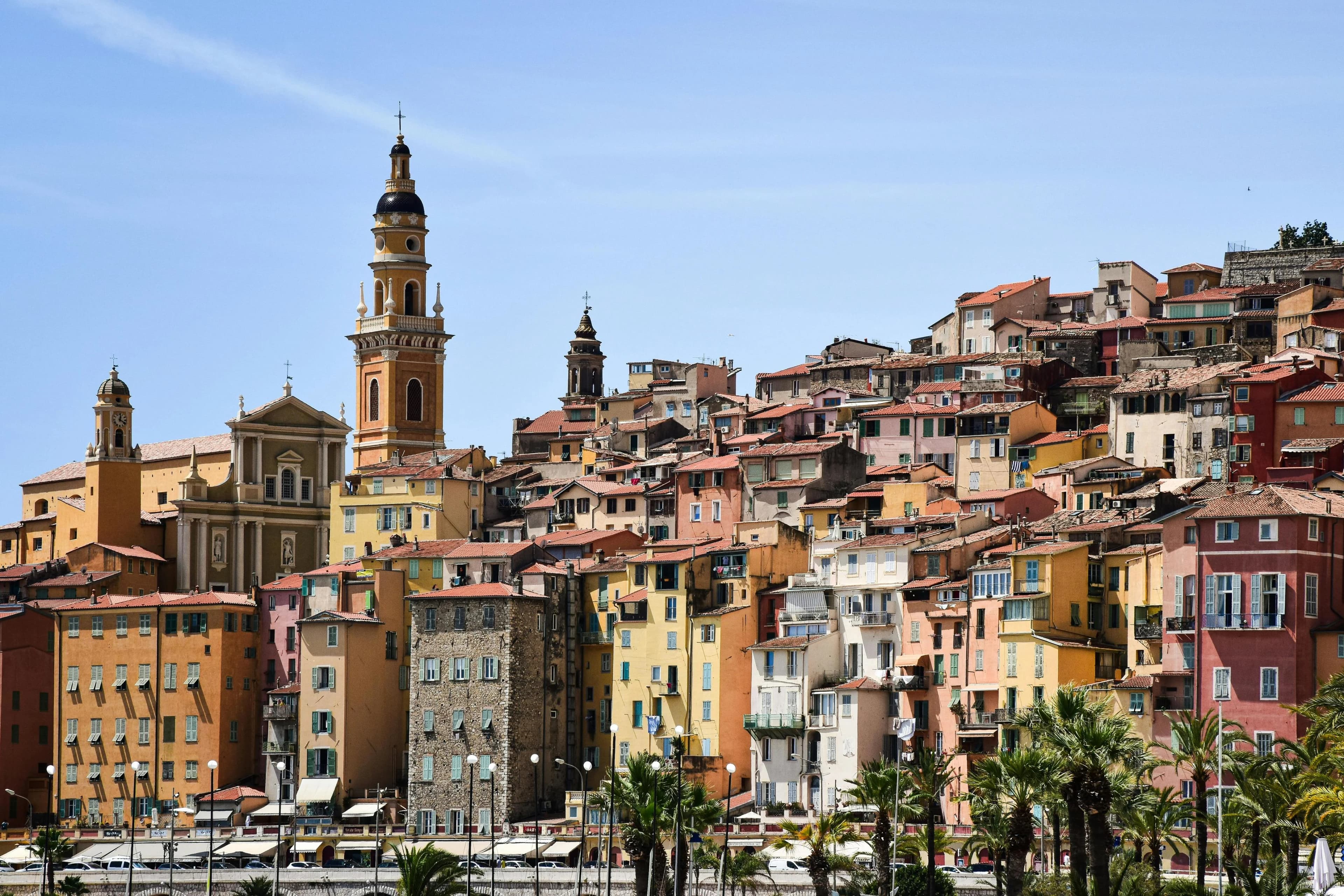 The vibrant, colorful buildings of Menton's Old Town climb up a hillside, with a magnificent bell tower and the sea in the foreground.