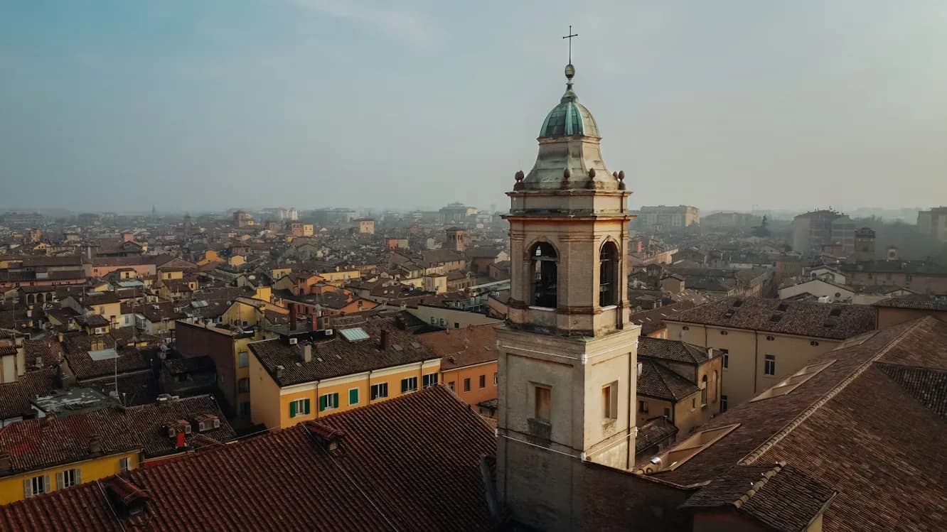 The bell tower of a historic church rises above the red-tiled rooftops of Modena, with a hazy sky in the background.