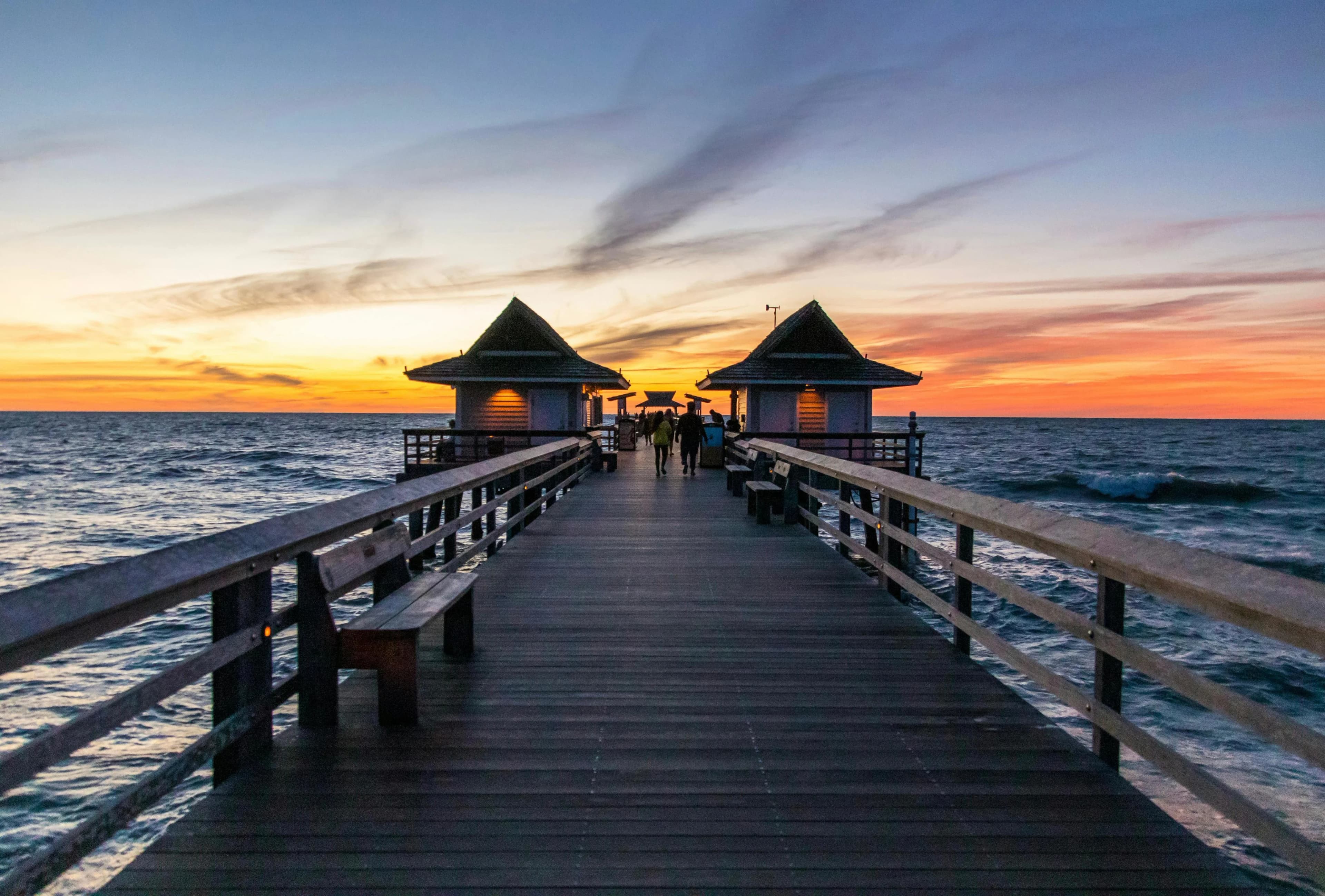 A long wooden pier with two small, covered huts stretches out over a calm sea at sunset.