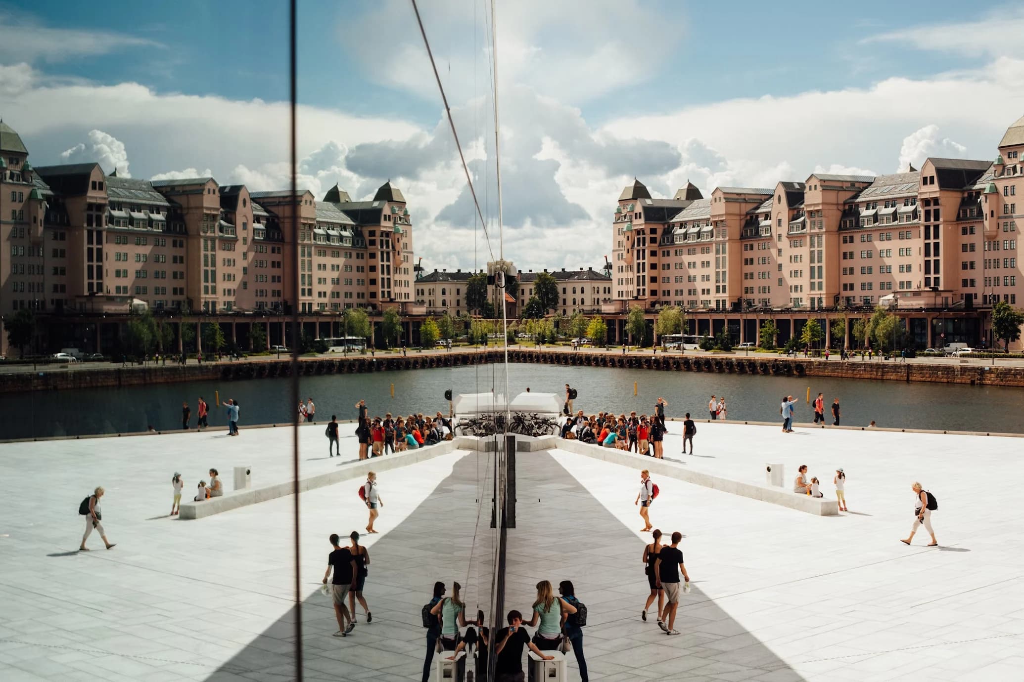 A modern, reflective building creates a surreal and artistic reflection of the city's waterfront, with people walking in the square below.