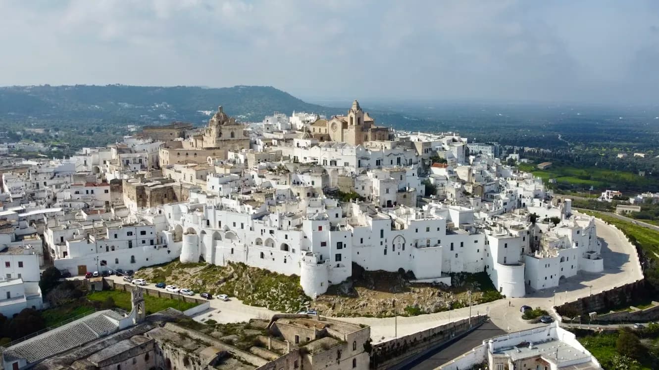 An aerial view captures the magnificent white city of Ostuni, perched on a hill and surrounded by green fields.