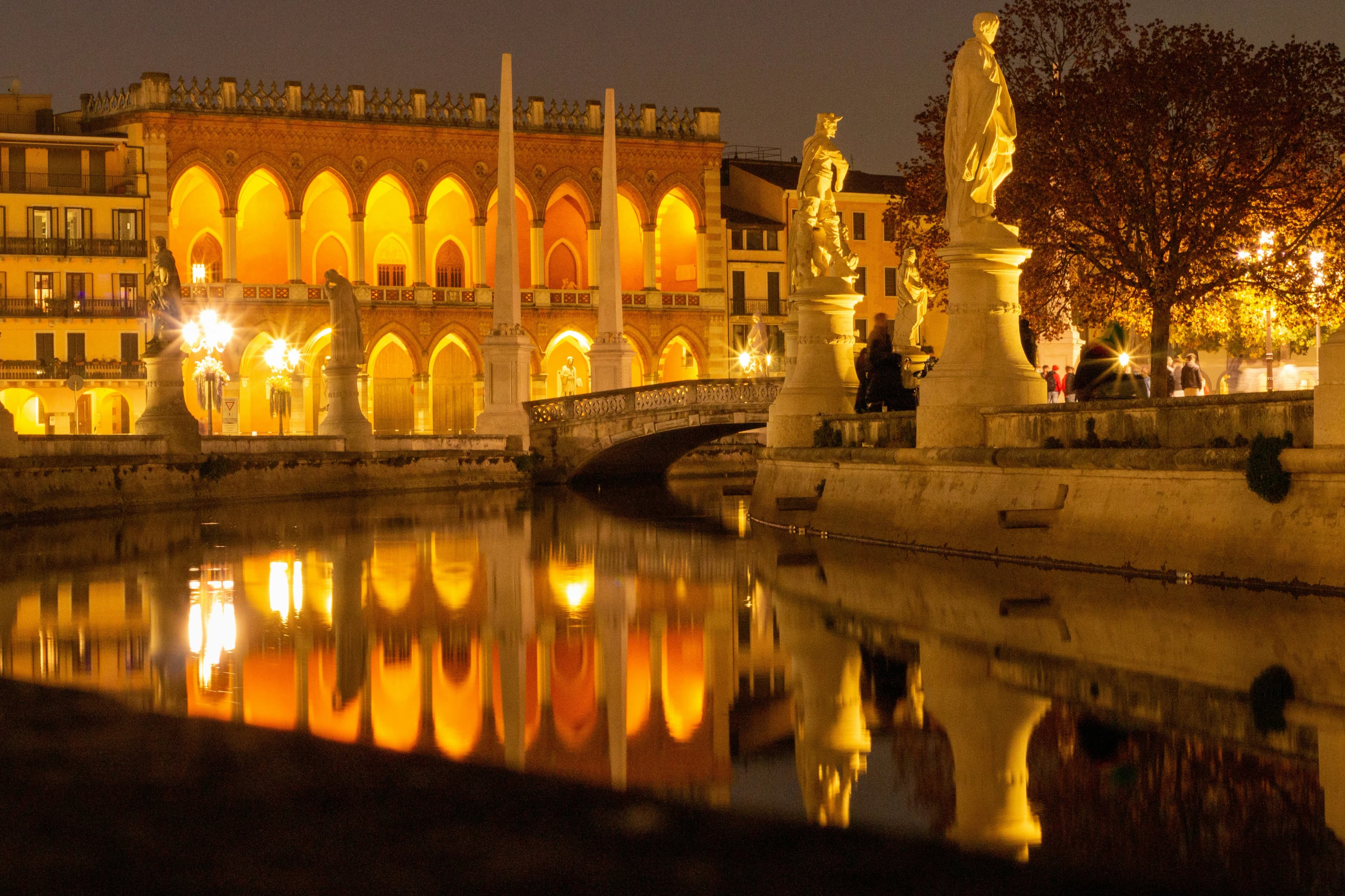 The illuminated arches of the Prato della Valle and the historic buildings of Padua are beautifully reflected on a calm river at night.