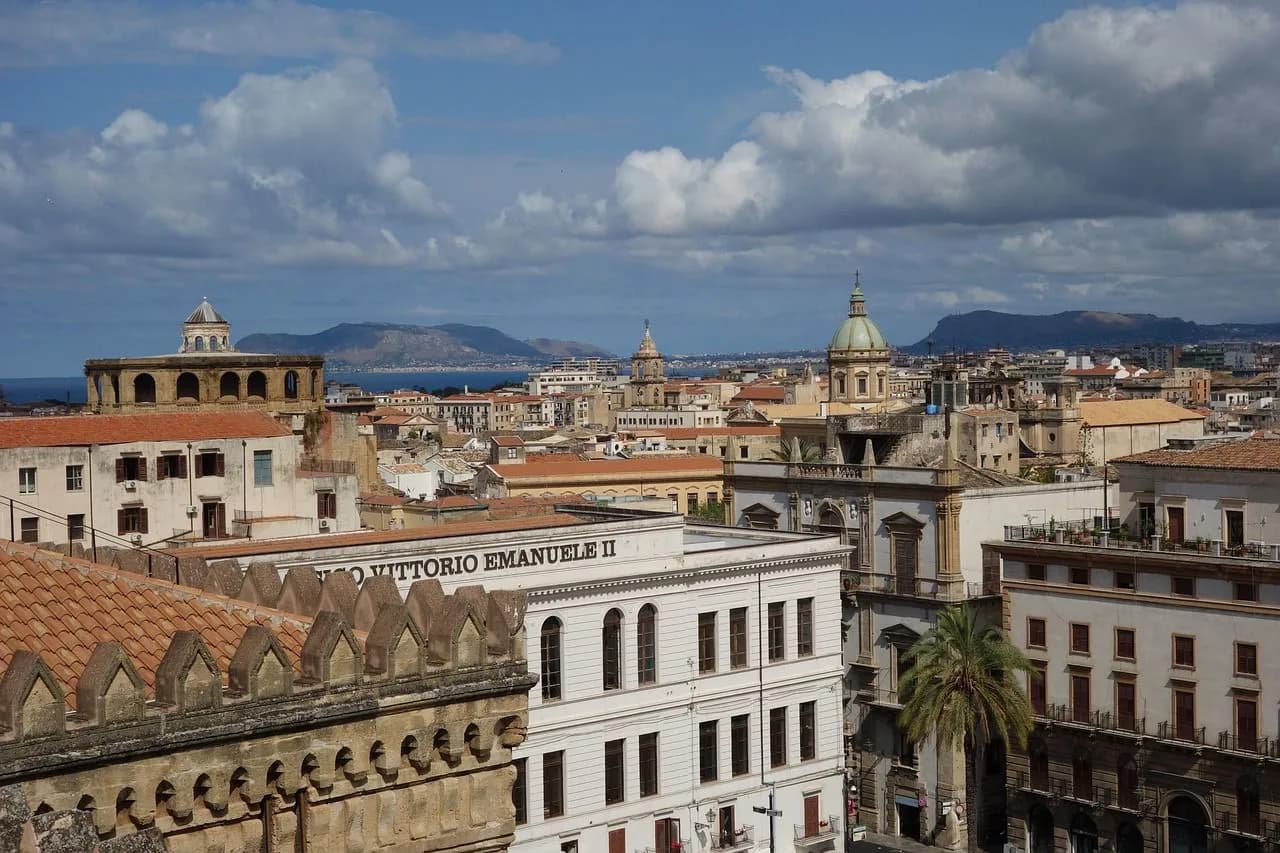 An aerial view captures the historic rooftops and domes of Palermo, with the sea and mountains in the distance.