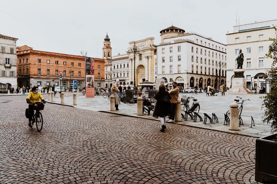 A bustling city square in Parma is filled with people, bicycles, and historic buildings with a clock tower and a statue.