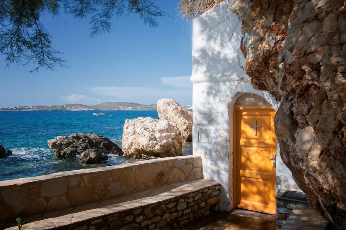 A traditional Greek chapel with a bright yellow door is nestled among large rocks on the edge of the clear blue sea.