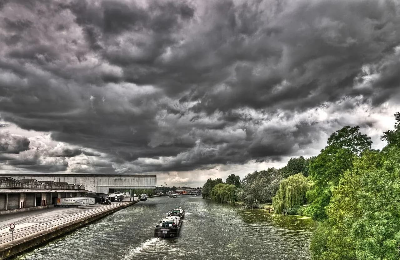 A large cargo boat travels down a river under a dramatic, cloudy sky, with a warehouse and green trees on the banks.