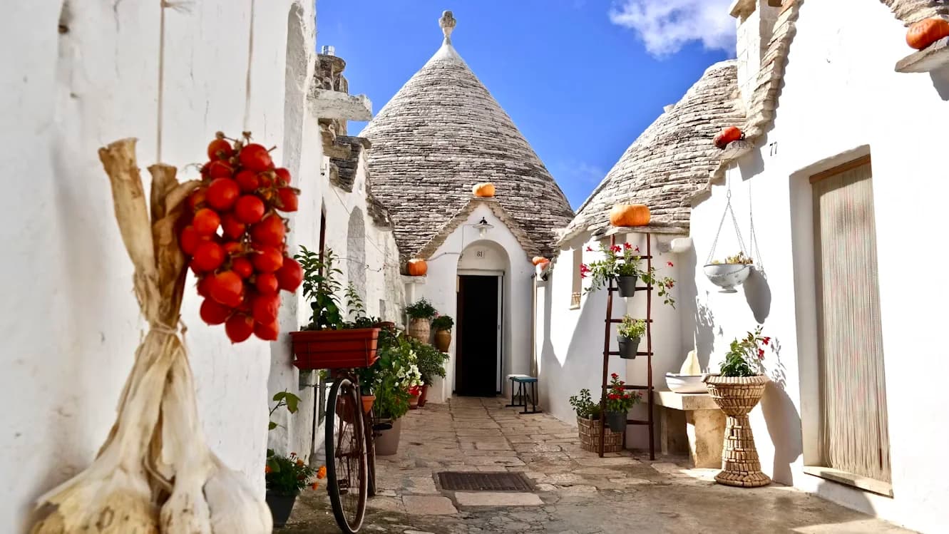 A traditional Trullo house, with its conical stone roof and white-washed walls, is nestled in a narrow, sun-drenched alleyway.