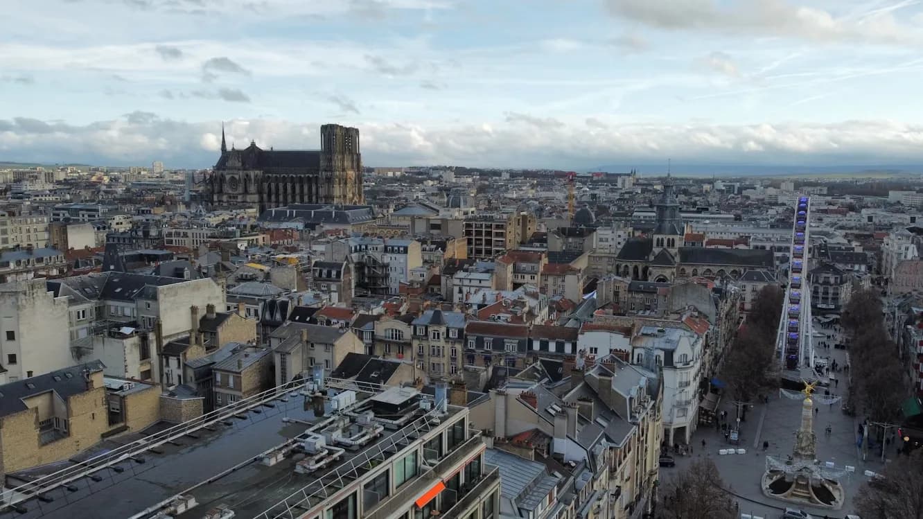 An aerial view captures the city of Reims, with the grand Reims Cathedral and a Ferris wheel in the distance.