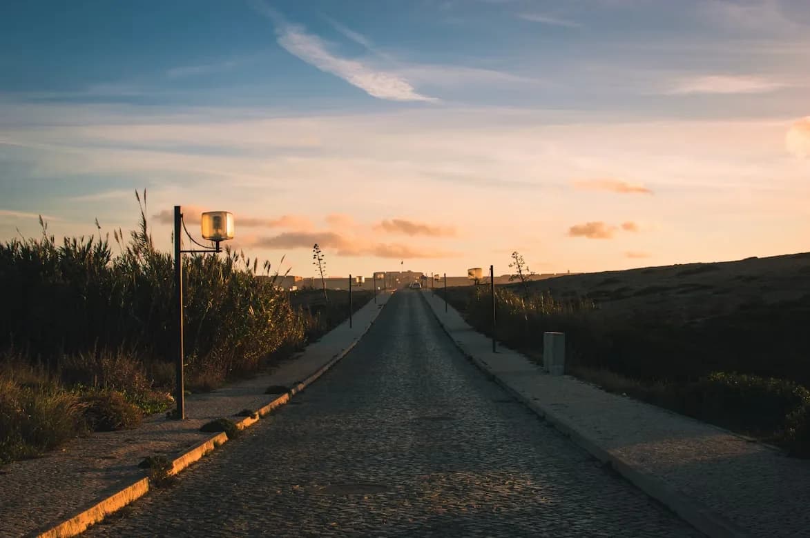 A wide cobblestone road with streetlights stretches into the distance, with the sun setting behind a hill.